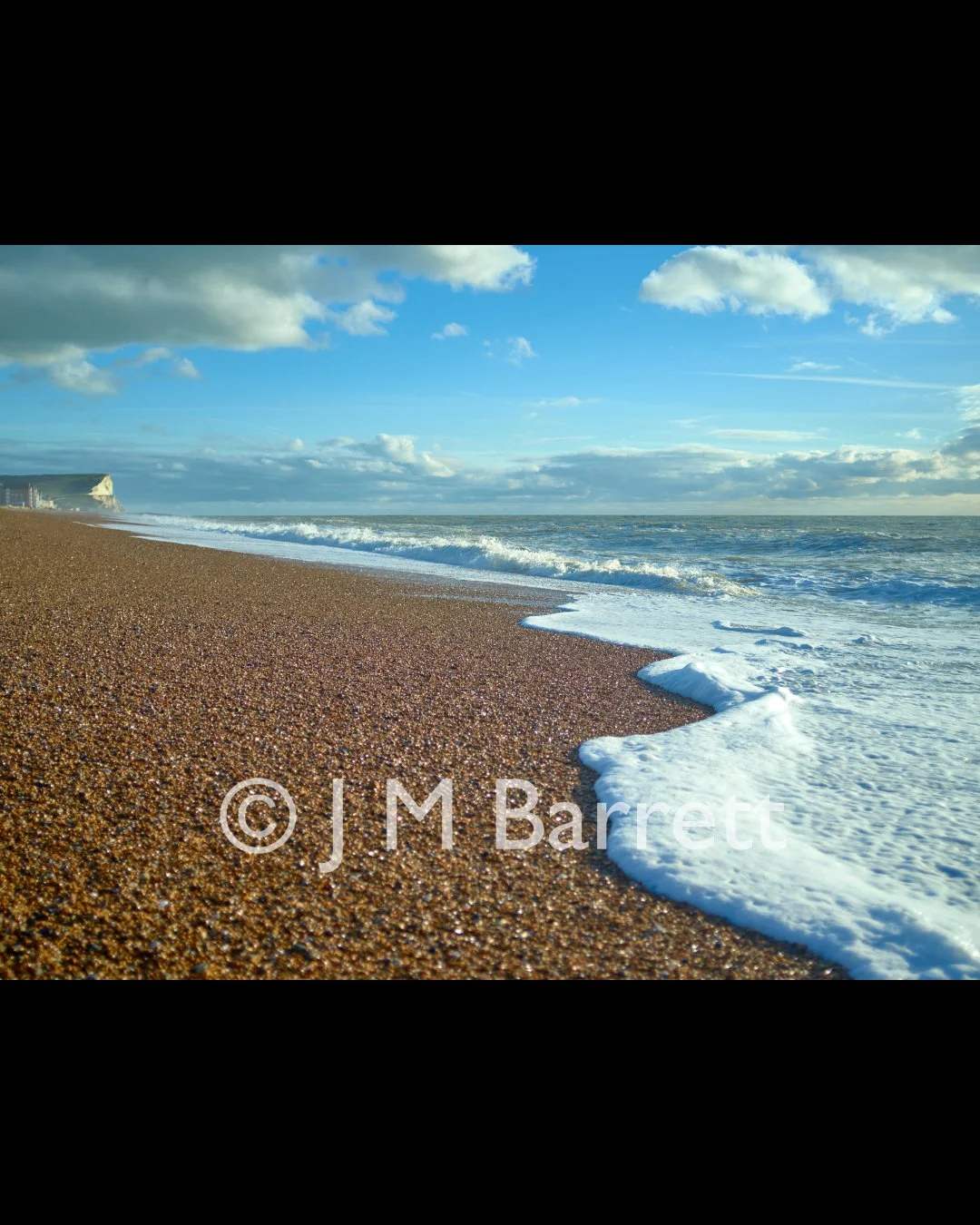 A sandy beach with small pebbles, ocean waves washing ashore, and a partly cloudy sky.