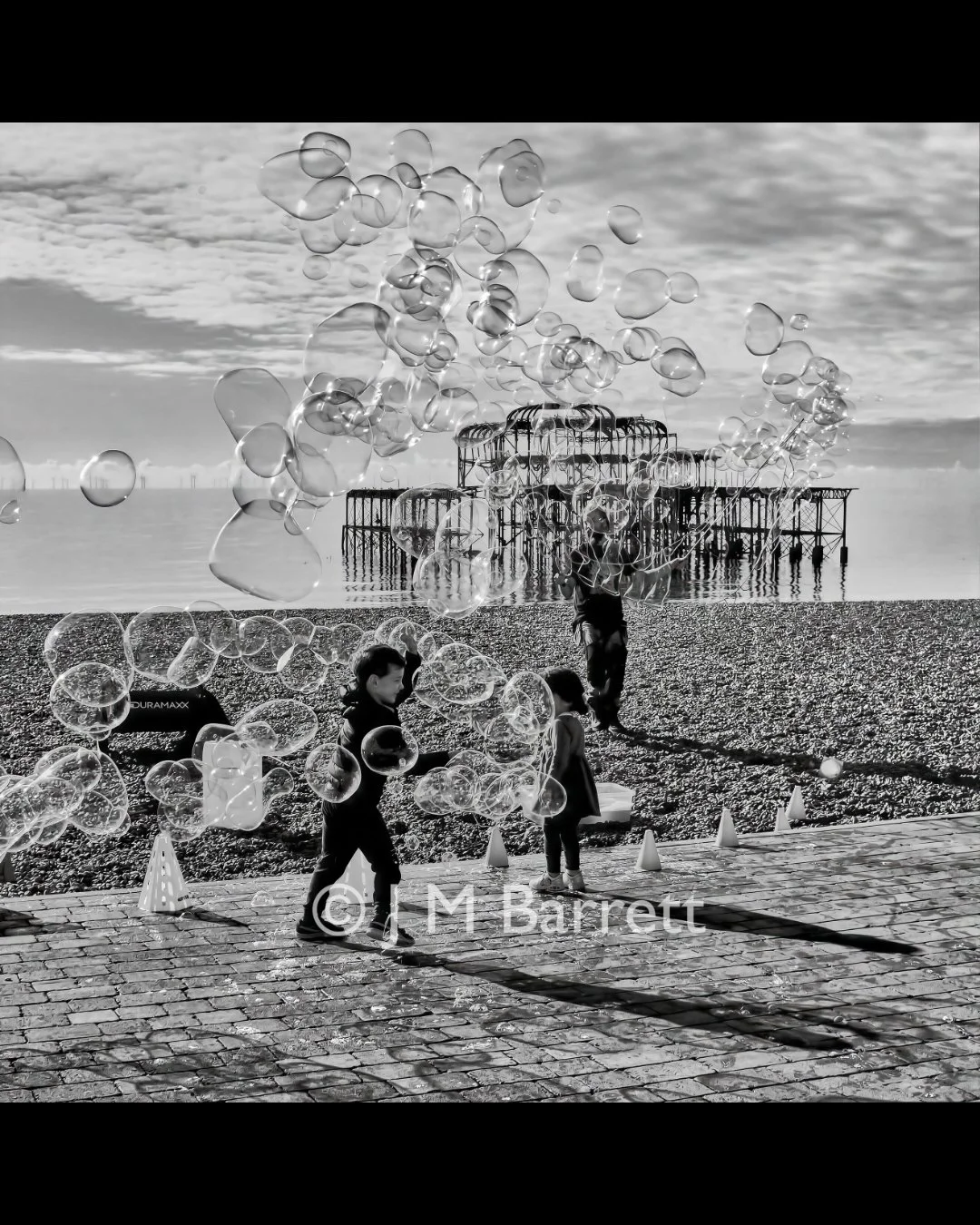 Children on a pebble beach playing with bubbles near a pier, with the ocean and cloudy sky in the background.