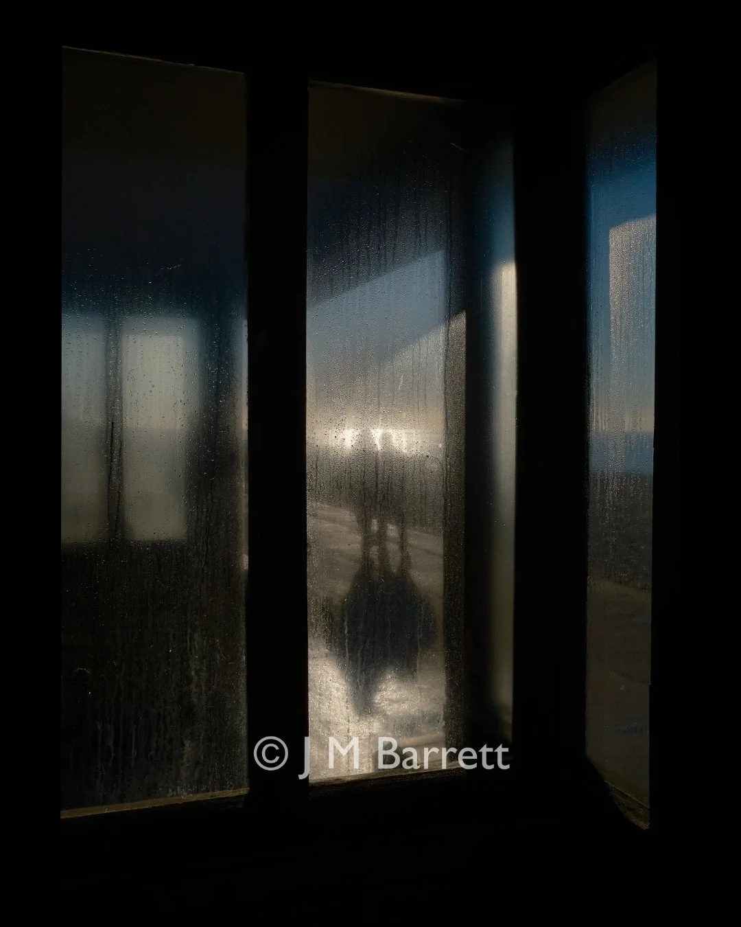 View through a rainy window with condensation, showing a silhouette of a person outdoors during sunset.