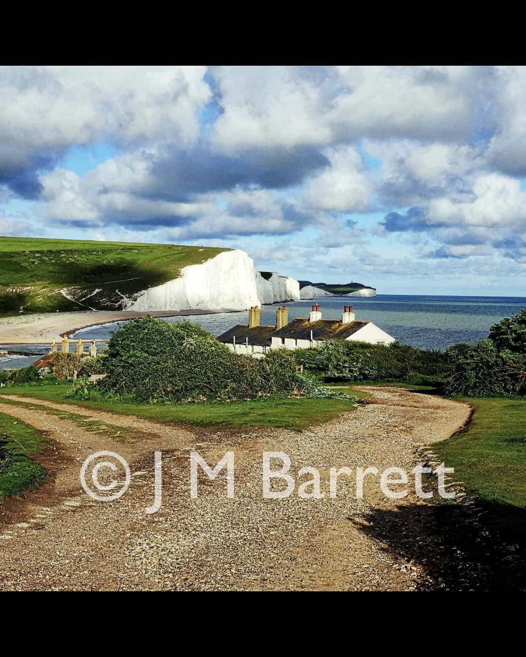 Dirt path leading to white cottages on green grass with chalk cliffs and ocean in background under cloudy sky.