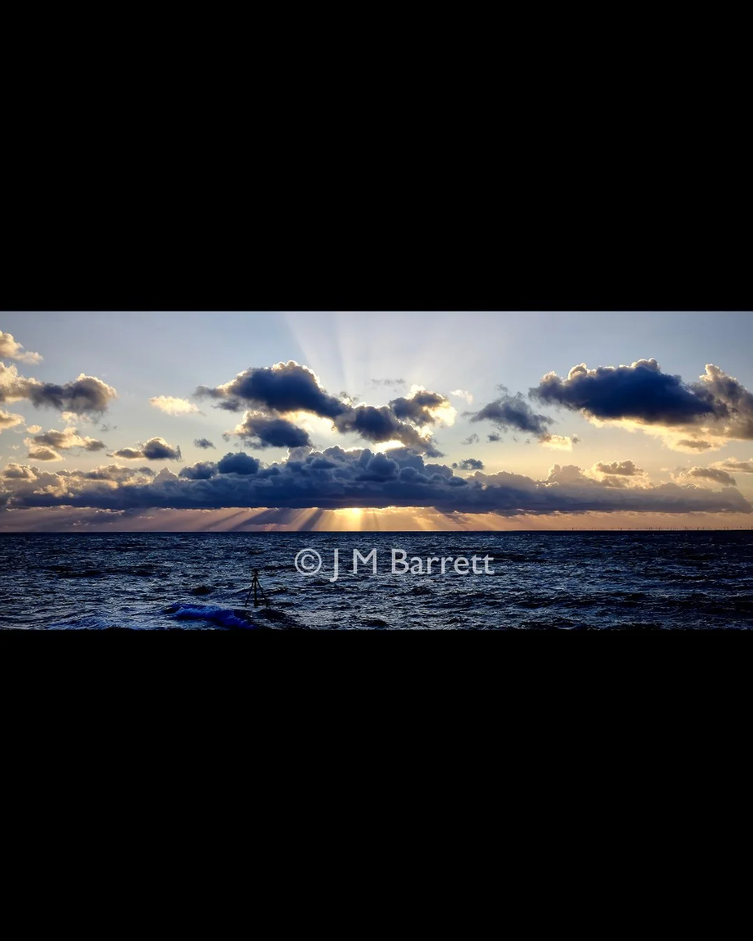 Sunset over ocean with partly cloudy sky, rays of sunlight breaking through clouds, photographer's tripod in water, copyright by J M Barrett.