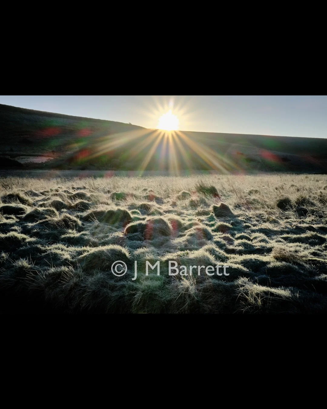 Sunrise over a foggy grass-covered landscape with a hill in the background, sunlight creating lens flare effects.