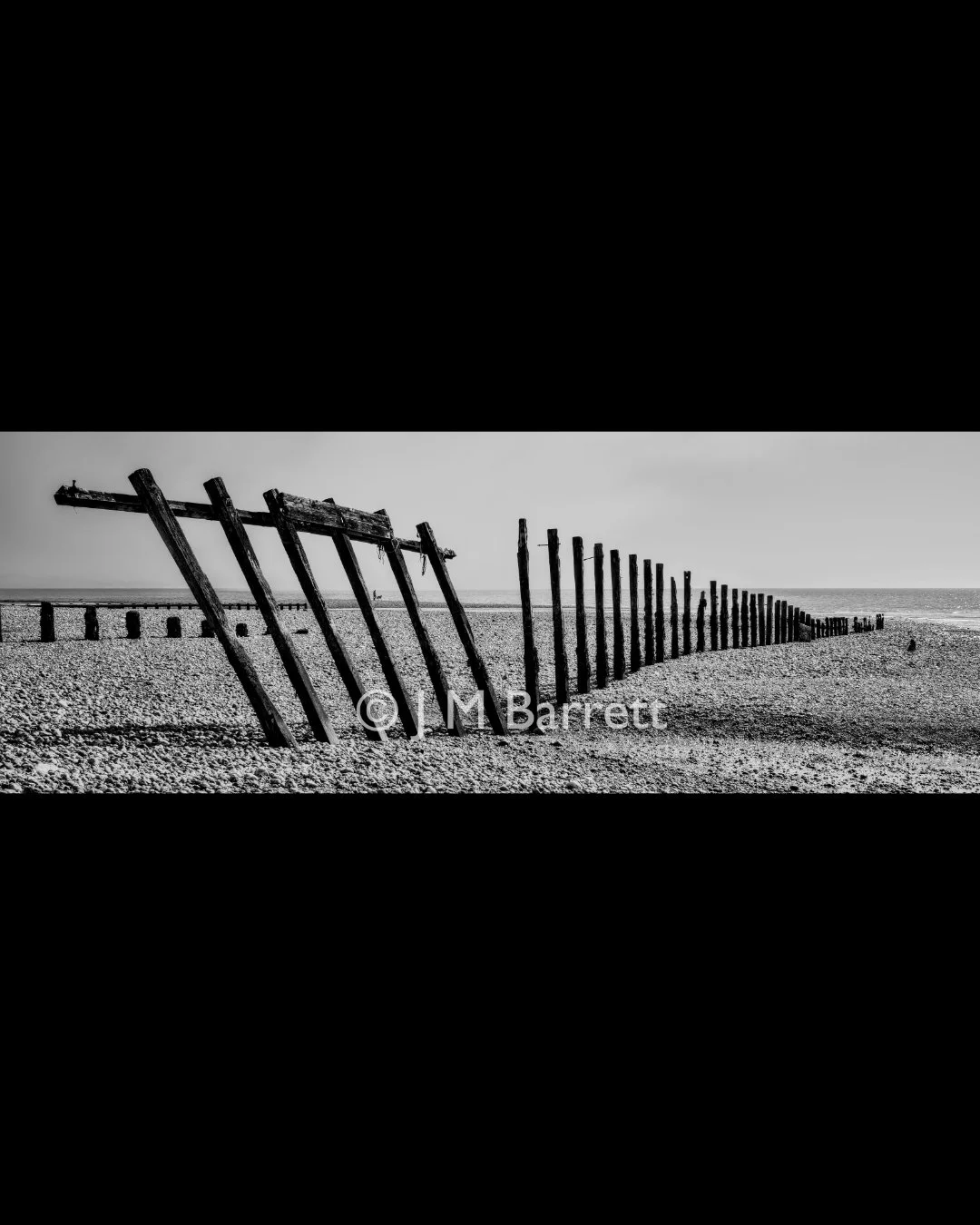 Black and white photo of a leaning wooden pier fencing on a beach, with the fence extending into the distance, over sand and near the water.