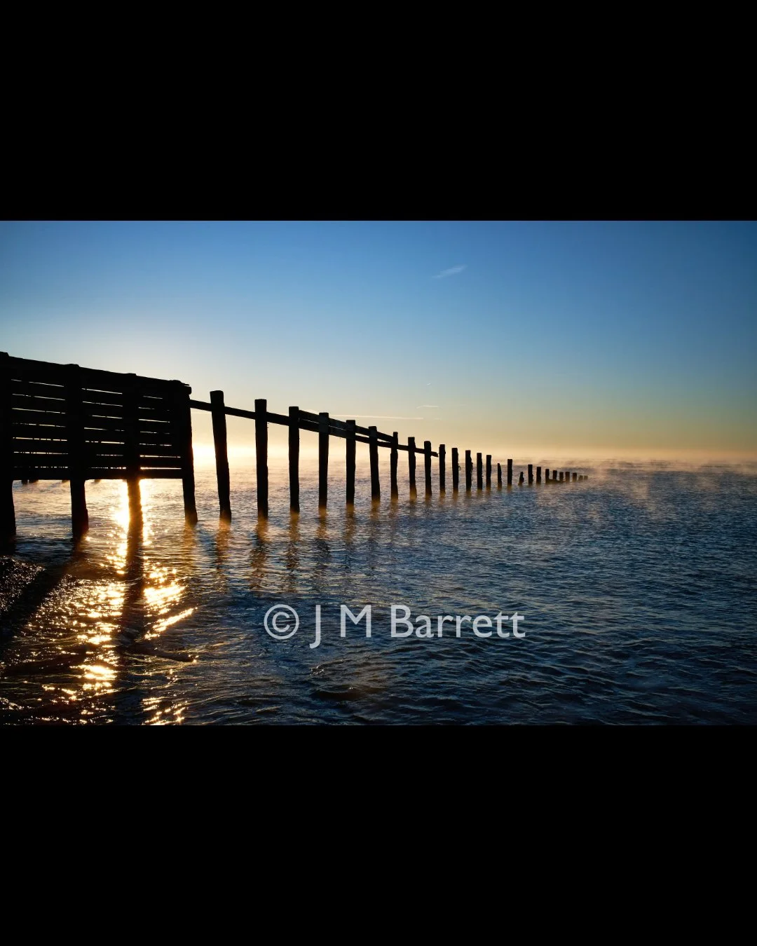 A wooden pier extends into the ocean at sunset, with the water reflecting the sky and the sun low on the horizon.