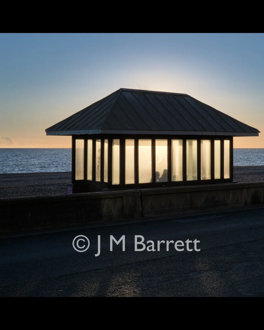 A small pavilion with a metal roof and glass walls is located on a beach at sunset, with the ocean in the background.