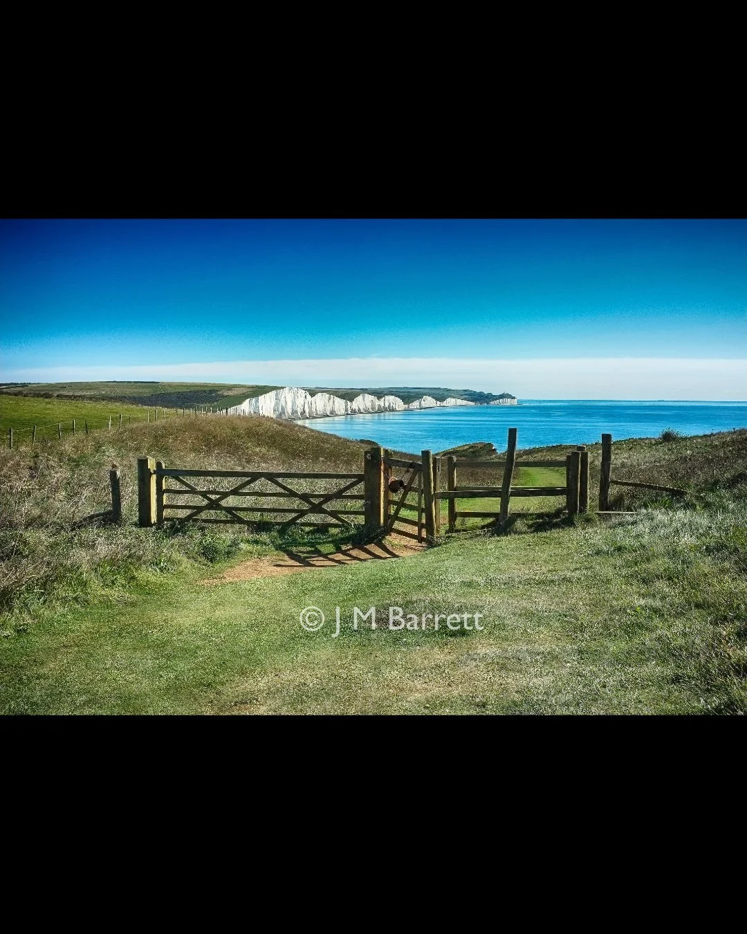 A grassy field with a wooden gate opening to a view of white chalk cliffs along the coast, blue sea, and clear sky.