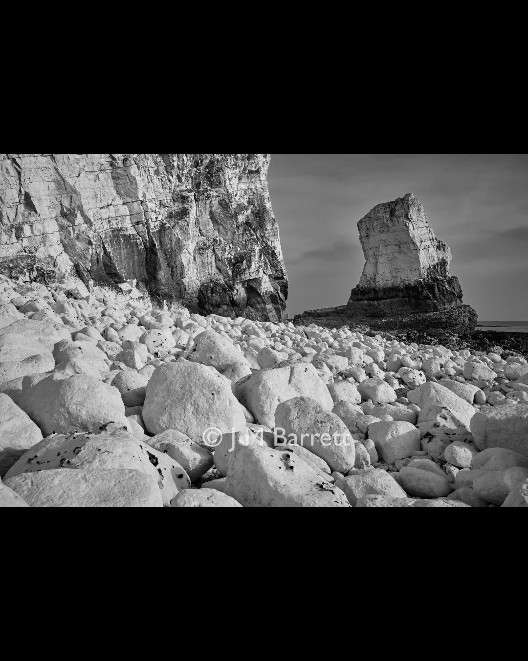 Black and white photograph of a rocky beach with large stones and a prominent white chalk cliff on the left and a large, isolated chalk rock formation in the sea to the right.