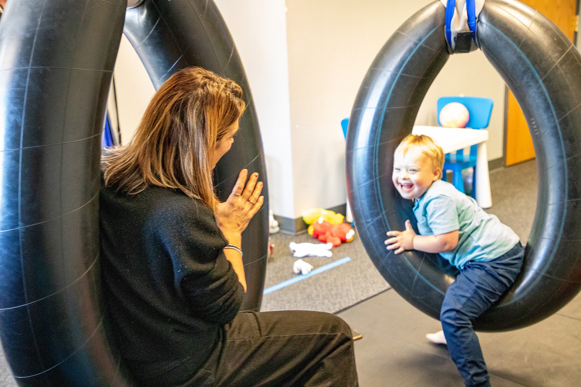 A woman and a young boy playing on large black inner tube swings in a playroom. The boy is smiling and kneeling inside the swing, while the woman is sitting nearby, watching and smiling.