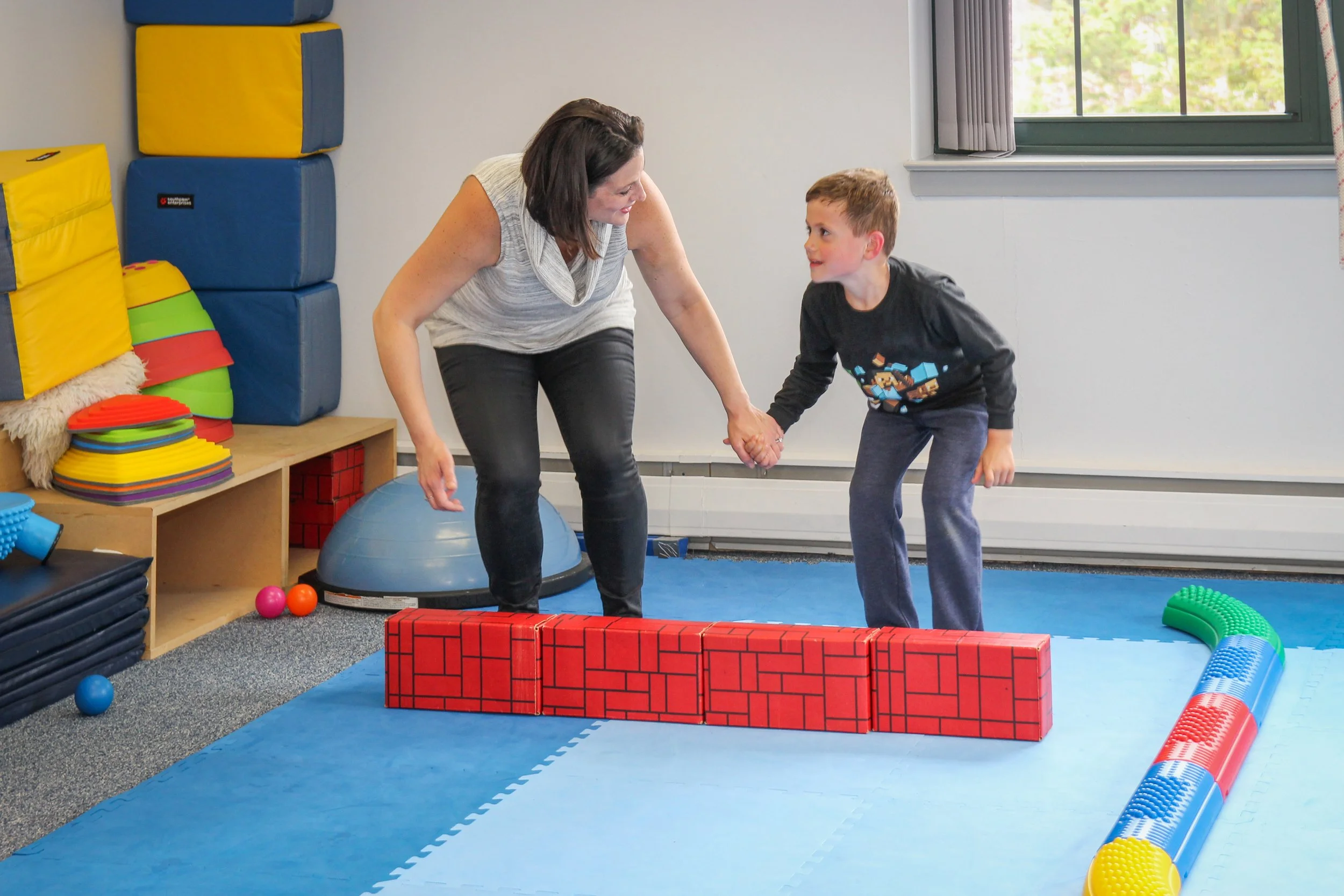 A woman and a boy are holding hands and smiling as they stand on a padded blue play mat in a room filled with soft play equipment and toys.