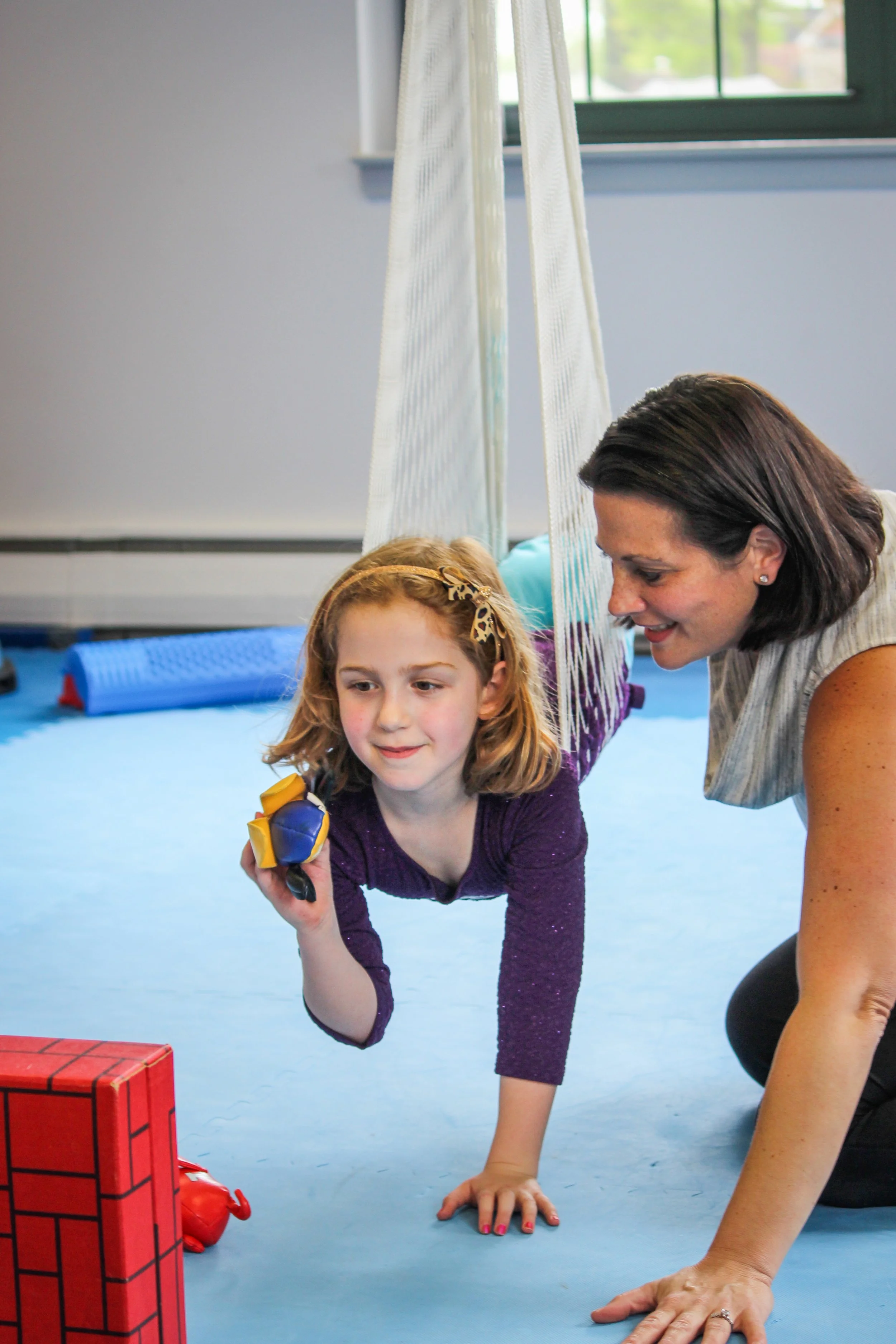 A girl doing a crawling exercise on the floor, supported by an adult woman, in a therapy or gym setting.