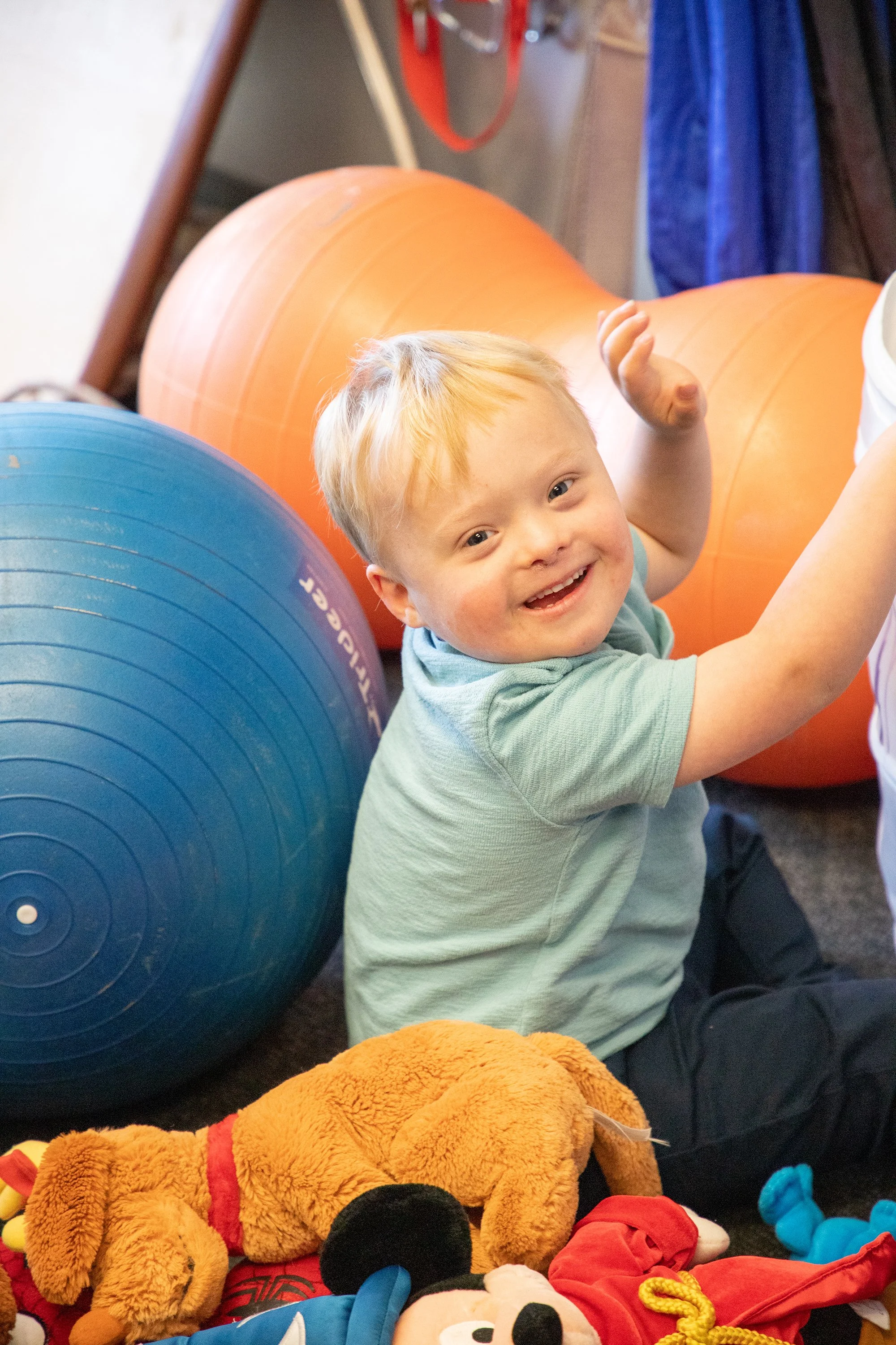 A young boy with blonde hair and a big smile sitting on the floor amidst toys and exercise balls, with large orange and blue exercise balls behind him.