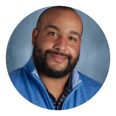 Headshot of a man with a beard, wearing a blue zip-up jacket over a checkered shirt, smiling against a painted blue background.