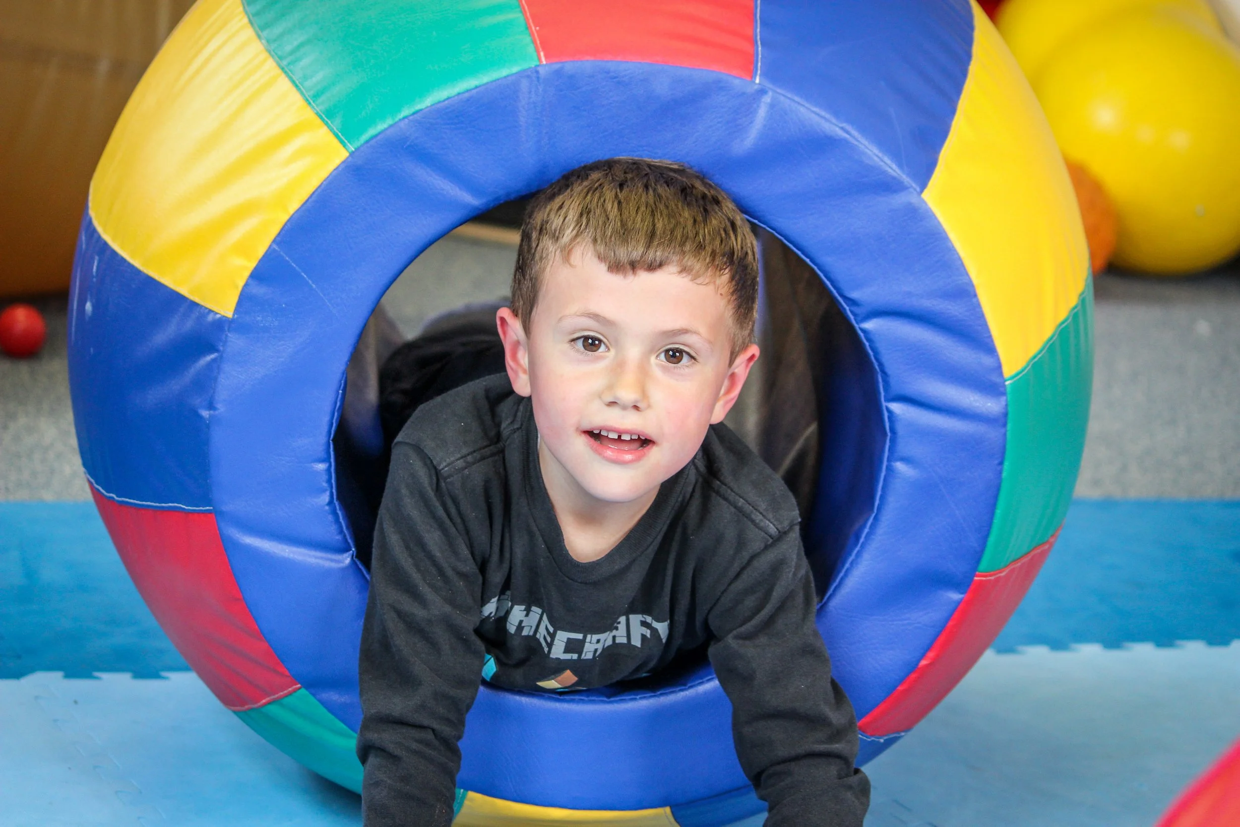 A young boy with short brown hair and a black shirt is crawling through a colorful, padded play tunnel at an indoor play area.