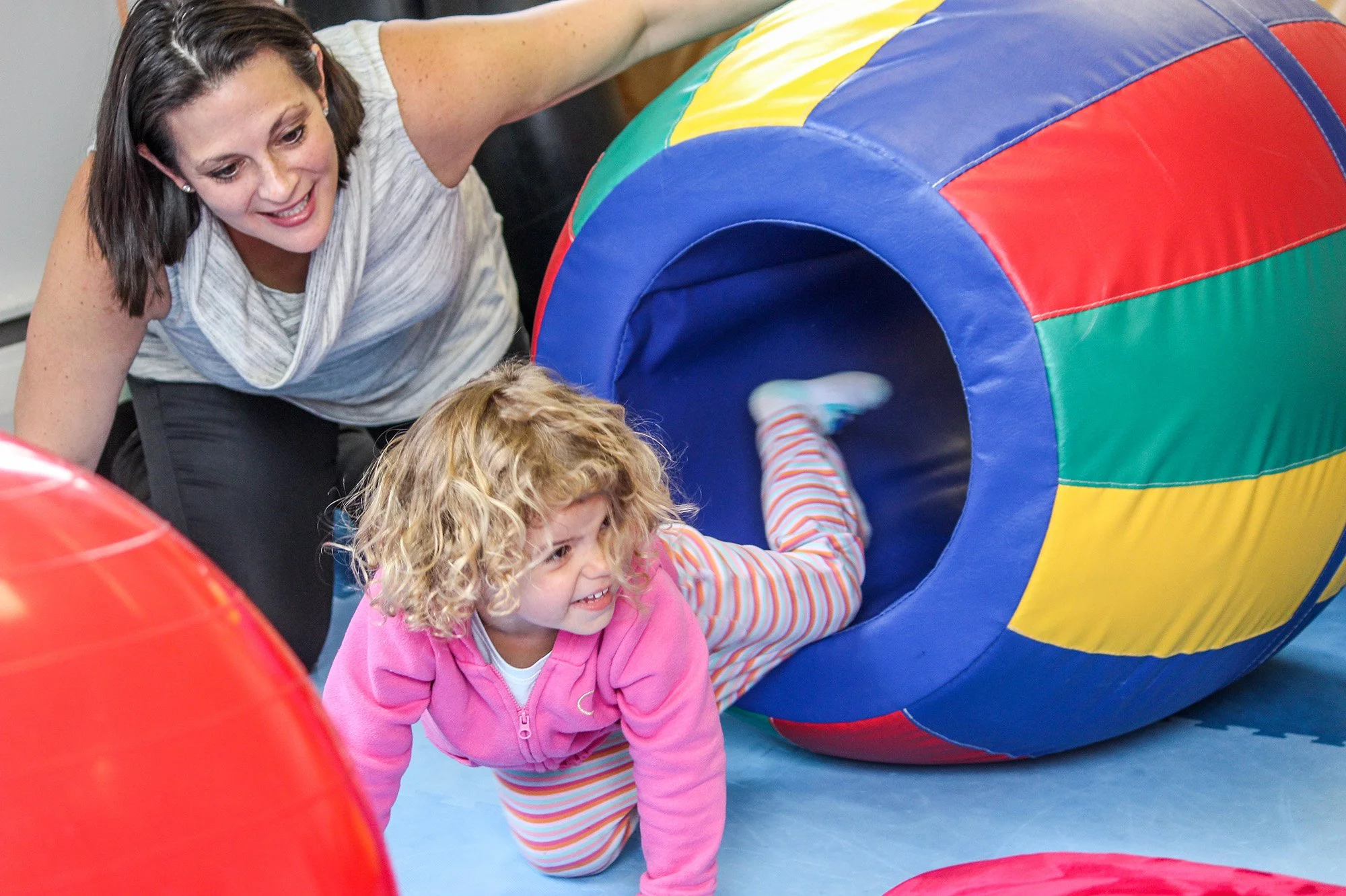 A young girl crawling through a colorful fabric tunnel with a woman supervising and smiling.