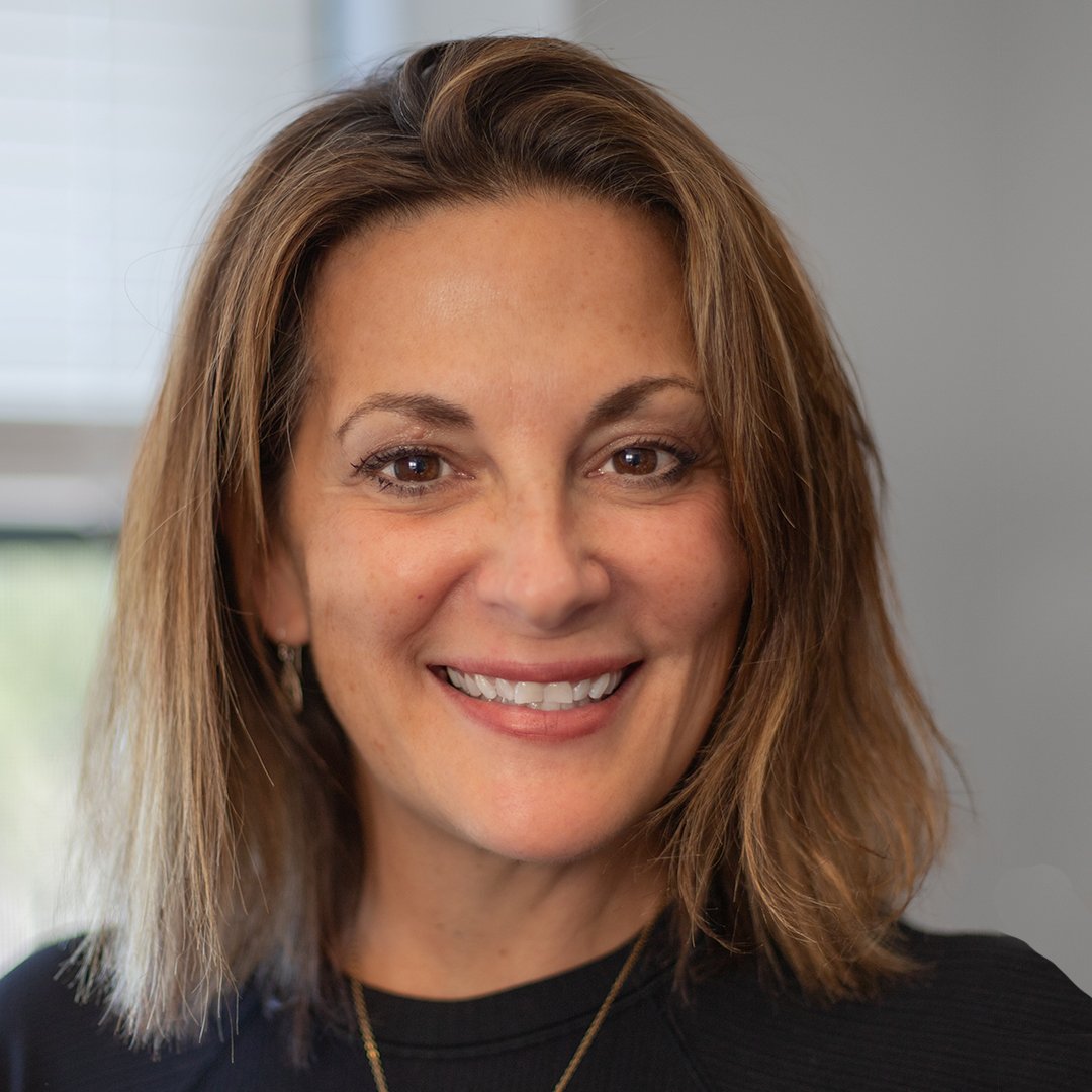 A woman with shoulder-length light brown hair, smiling, wearing a black top and earrings, standing indoors with a window in the background.