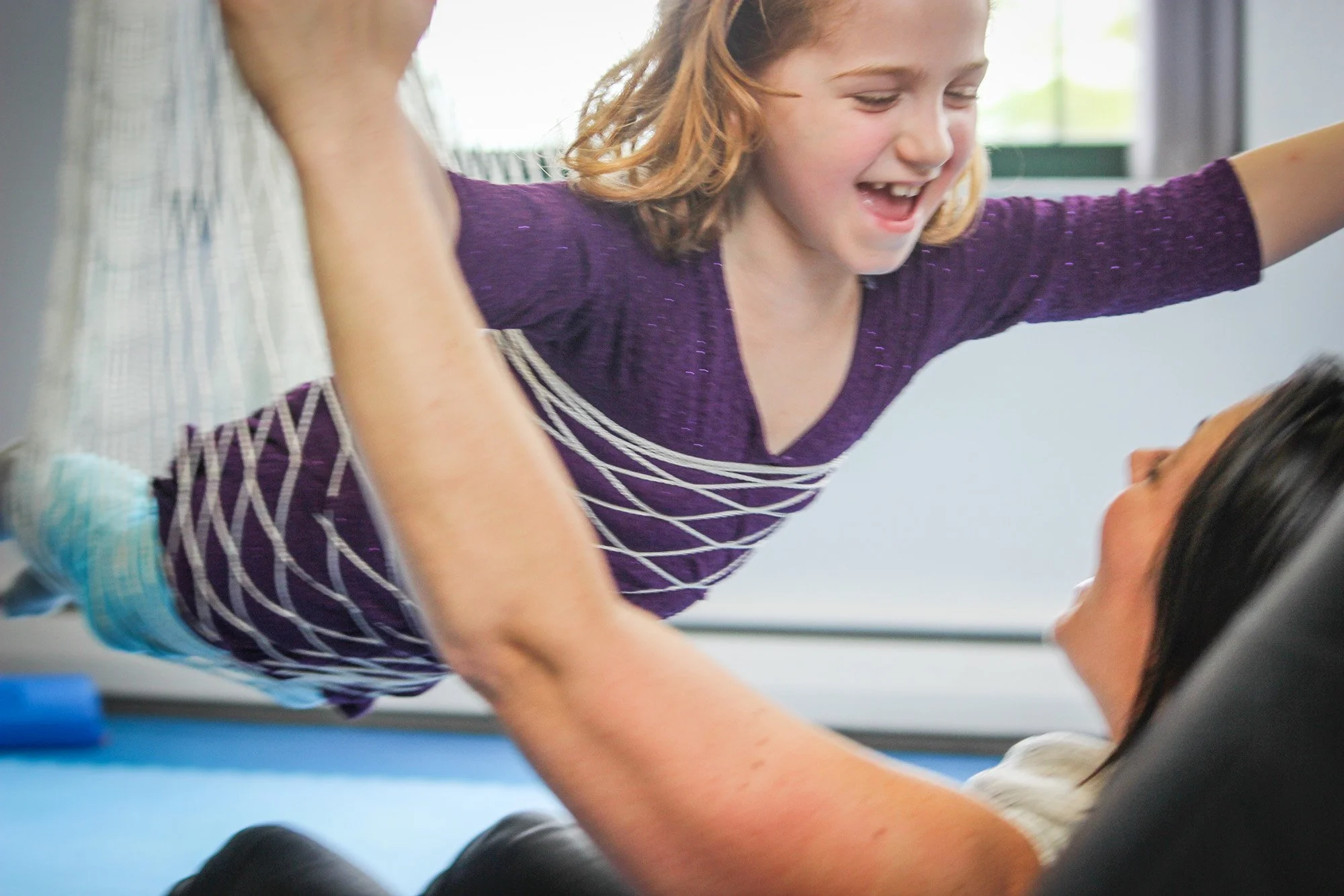 A young girl with red hair and a smiling face, wearing a purple shirt with white-and-purple checkered shorts, is laughing and playfully balancing on her mother, who is lying on the floor.