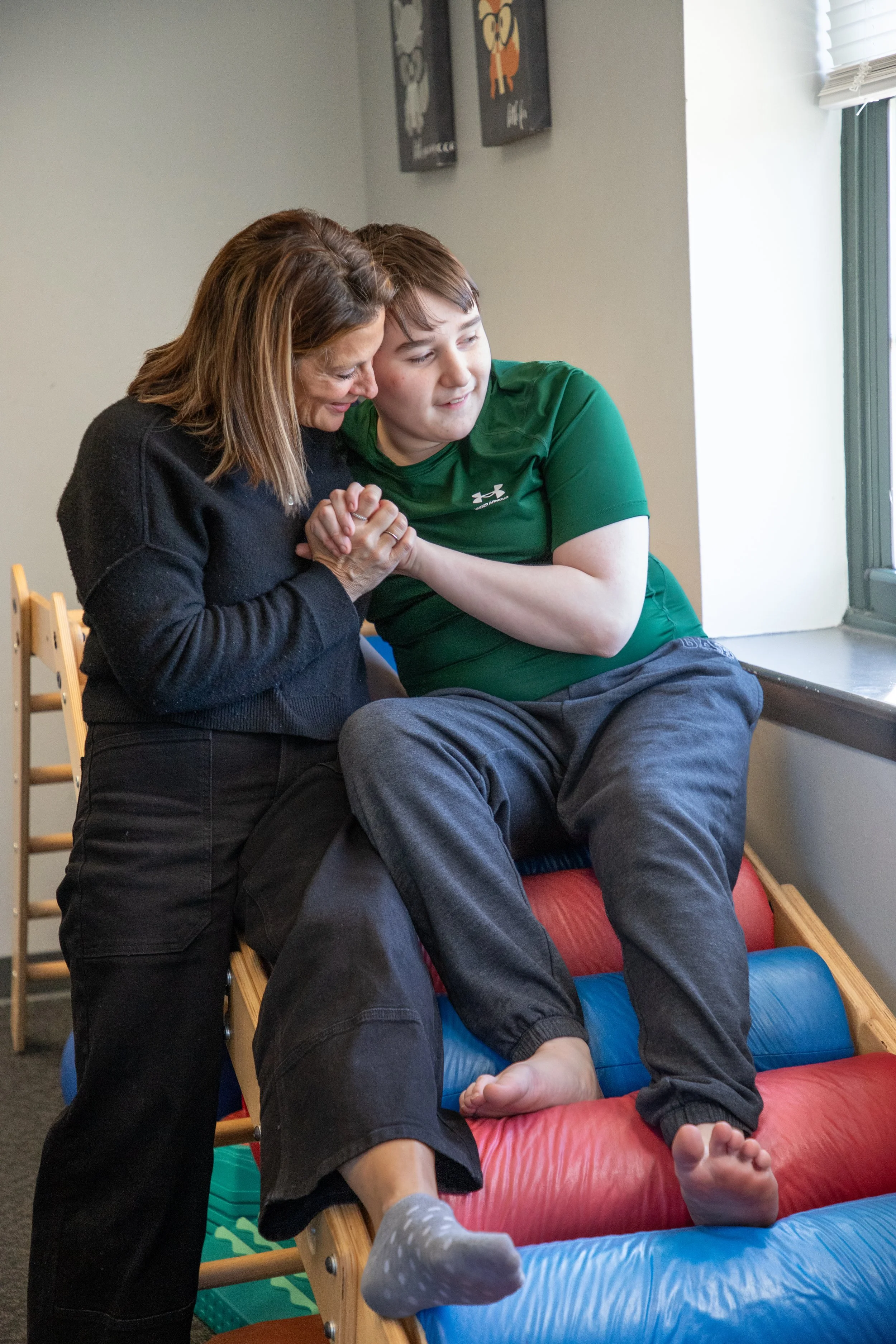 A woman and a boy are engaging in a therapy activity with their hands clasped together, the boy sitting on a padded inclined surface near a window.