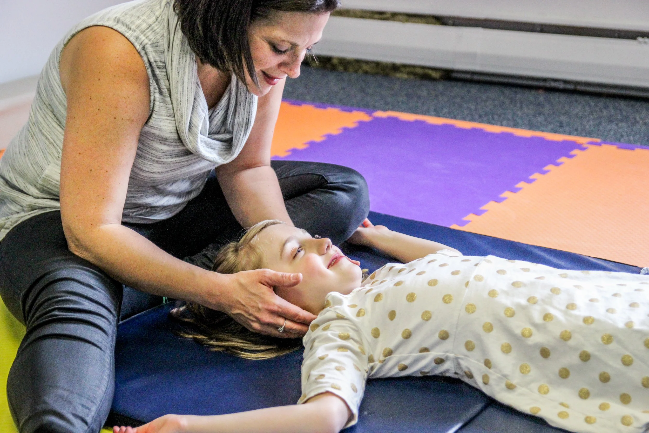 A woman placing her hand on a young girl's neck while lying on a mat during a children's chiropractic treatment.