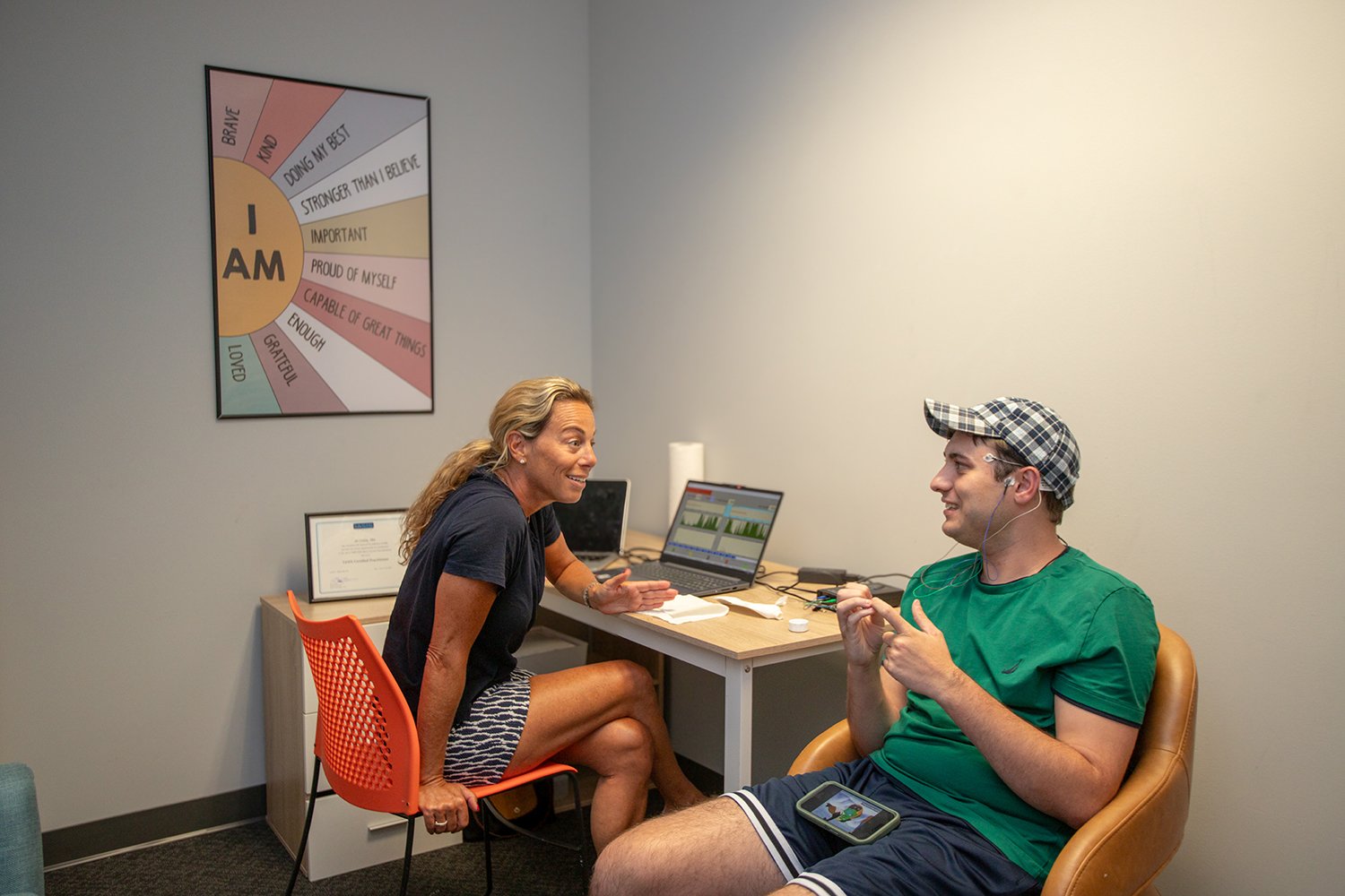 A woman and a man are having a conversation in a small office. The woman is sitting on an orange chair wearing a black shirt and striped shorts. The man is sitting in a tan chair wearing a green t-shirt, striped shorts, and a checkered cap, listening to music on earbuds. There are two laptops and some papers on the desk between them. A colorful motivational poster hangs on the wall behind the woman.