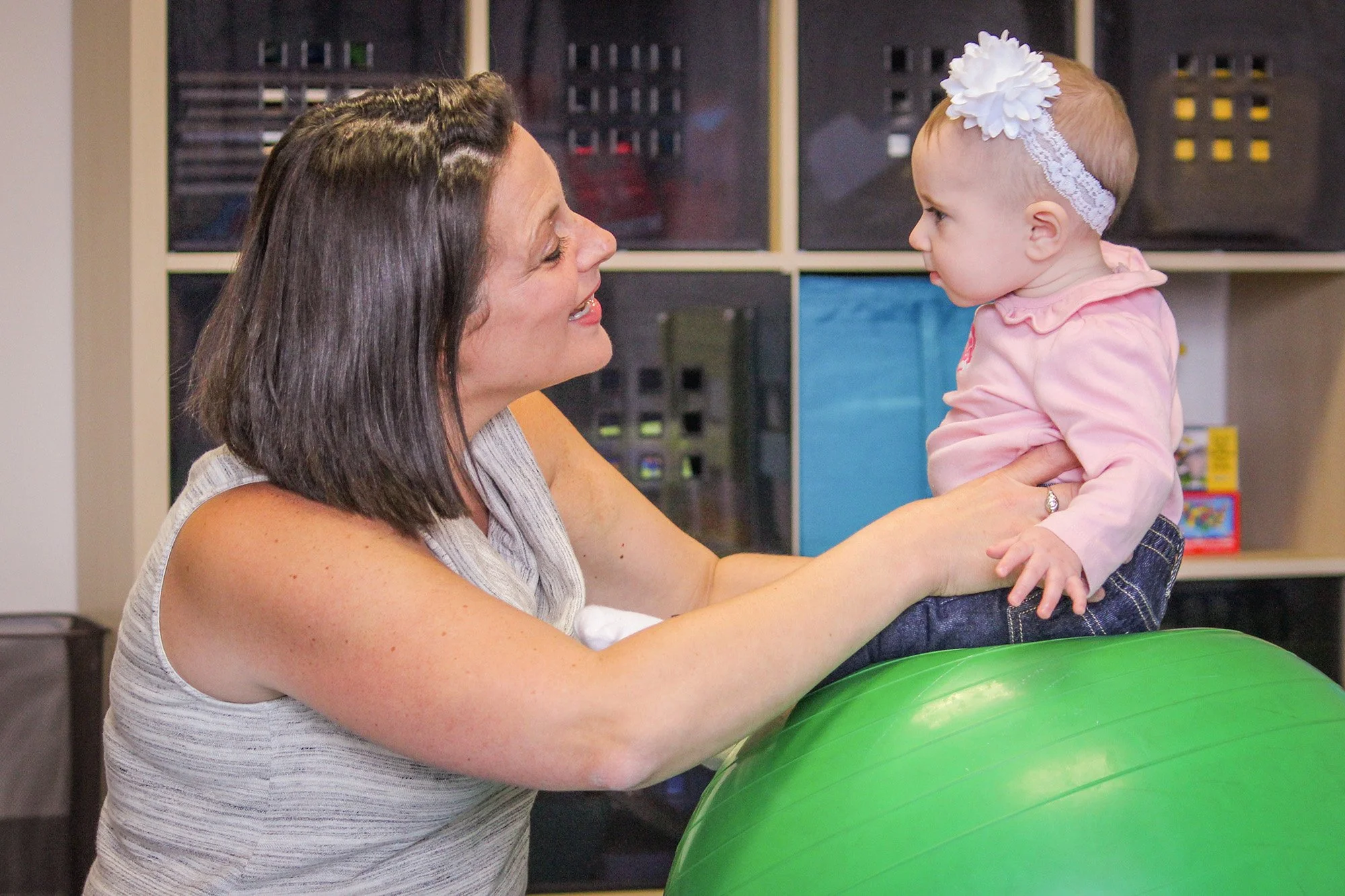 A woman holding a baby girl on her lap, both smiling and looking at each other. The woman is wearing a sleeveless top, and the baby is wearing a pink jacket and a white headband with a large flower. They are in a room with shelves and storage baskets in the background.