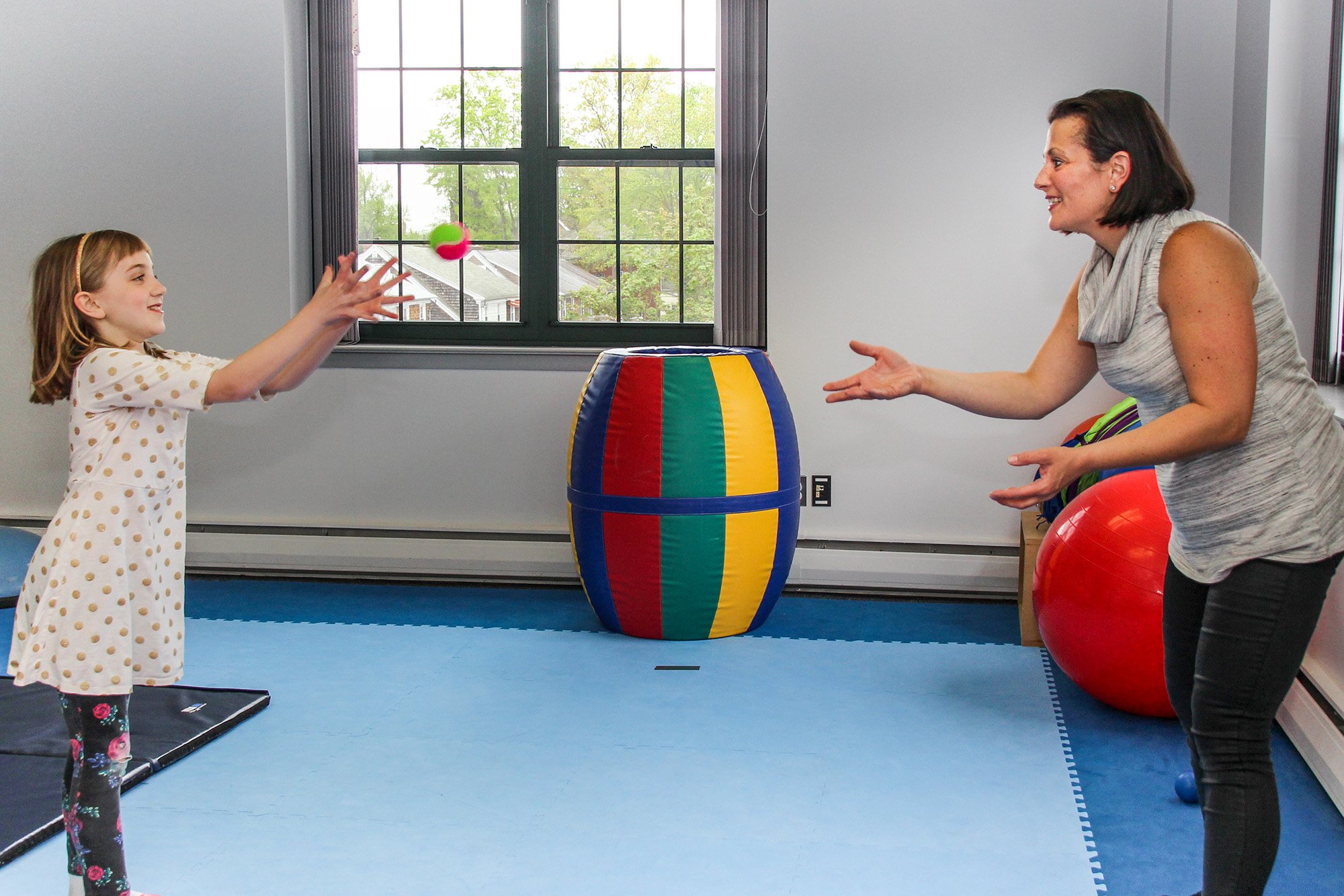 A young girl and an adult woman playing with a ball and a colorful barrel in a therapy or playroom with large windows and blue rubber flooring.