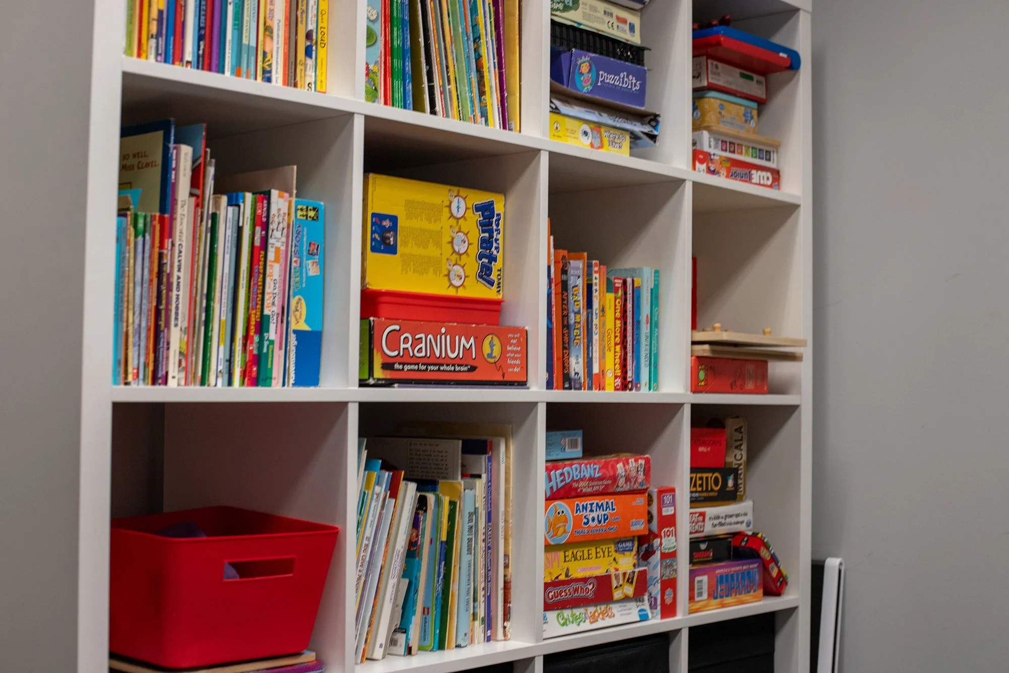 A white bookshelf filled with children's books, board games, and puzzles, with a red storage bin on the bottom left shelf and plain gray wall on the right side.