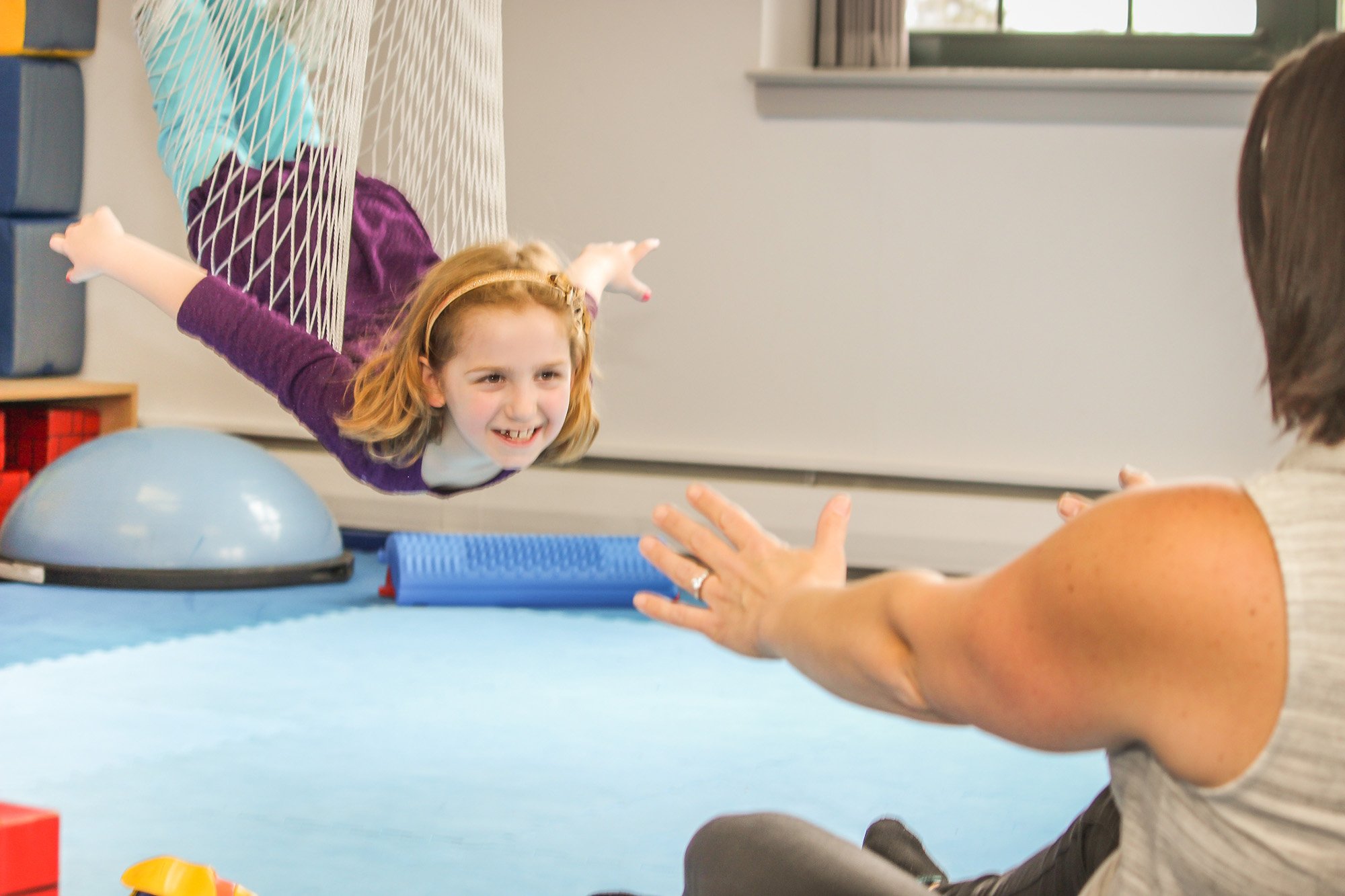 A young girl is flying in a crochet hammock over a padded gymnastics mat, reaching towards a woman sitting on the ground with her hand extended.