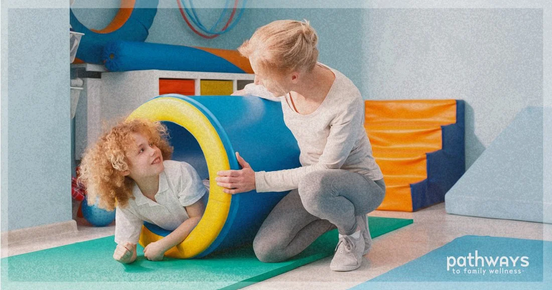 Child with curly hair crawling out of a blue and yellow foam tunnel with a caregiver nearby in a playroom.