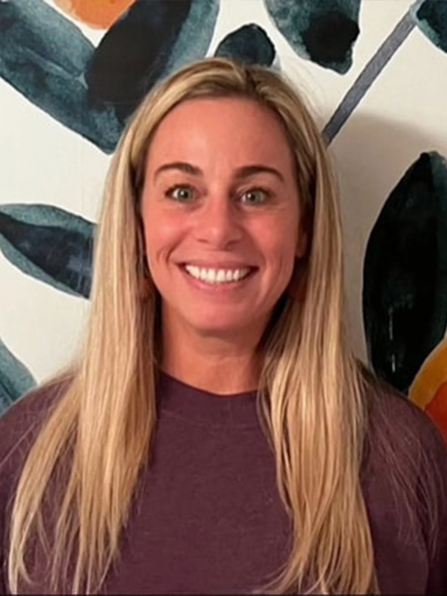 Smiling woman with long blonde hair standing in front of a leafy artwork.