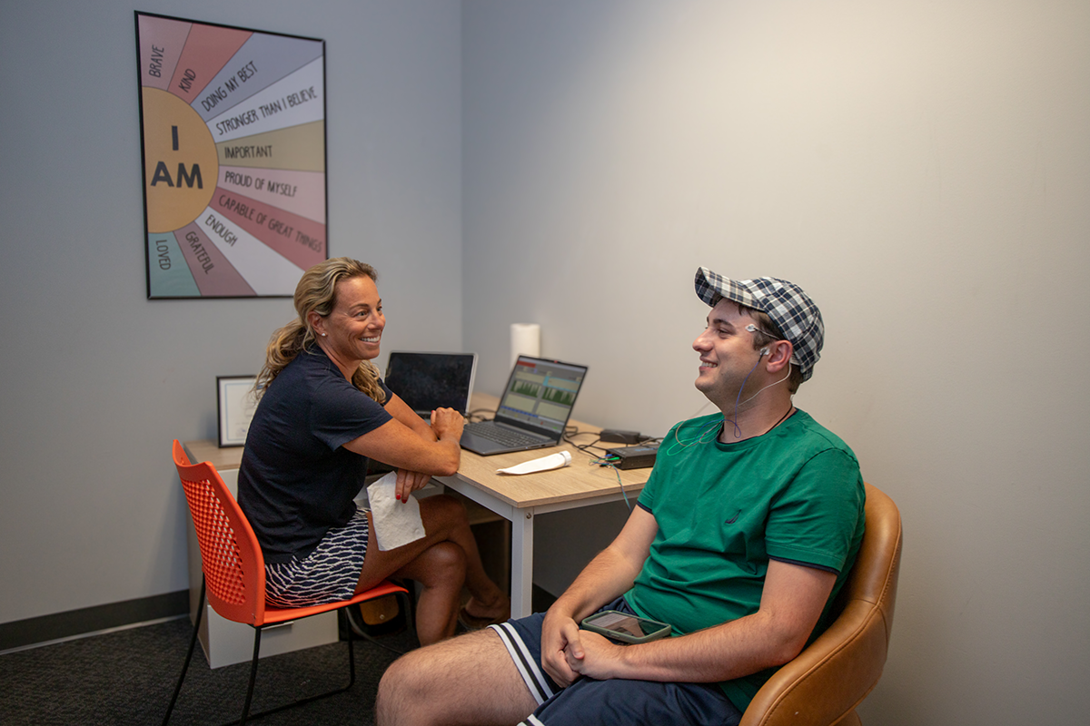 A woman and a man sitting in a therapy or counseling session, smiling and talking. The woman has a laptop in front of her, and the man is sitting in a chair holding a phone. A motivational poster is on the wall behind them.