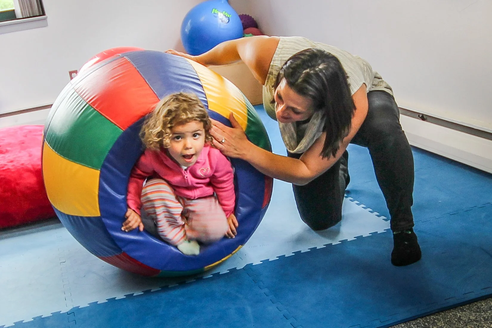 A young girl with curly blonde hair sitting inside a large, colorful play ball, with a woman beside her on her knees, holding the ball. The girl has a surprised or excited expression. The scene takes place on a blue foam mat in an indoor play area.