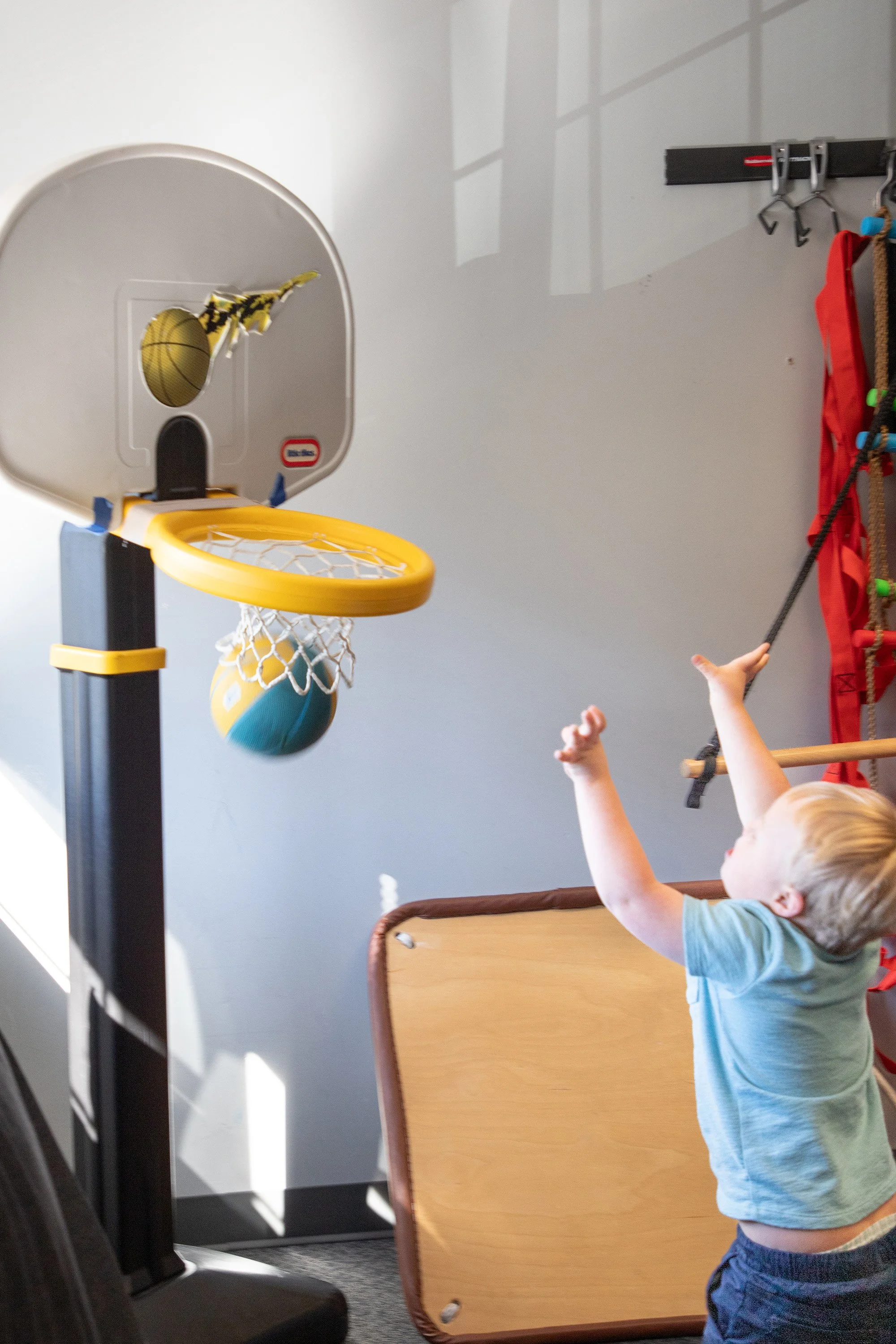 A young boy playing basketball indoors, shooting a ball towards a small yellow hoop attached to a mini basketball set, with a padded brown mat on the floor nearby.