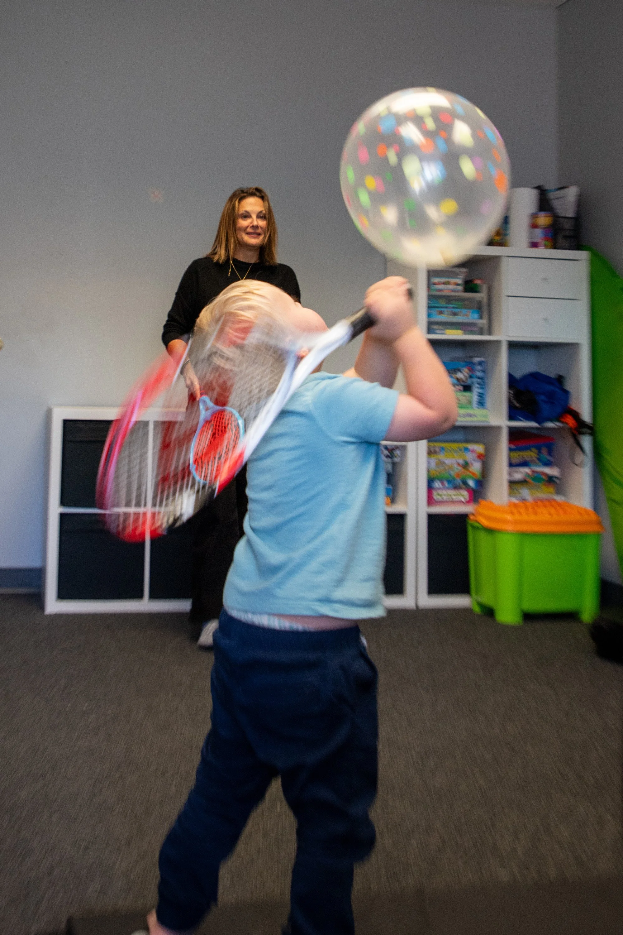 A child hitting a colorful helium balloon with a tennis racket, while an adult watches in a room with shelves filled with games and books.