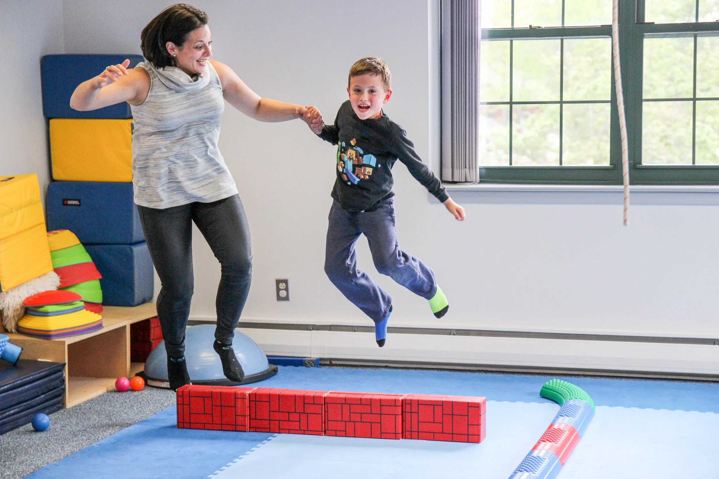 A woman and a young boy jumping on a padded play area inside a room, with the woman holding the boy's hand. The room has colorful padded blocks and a window in the background.