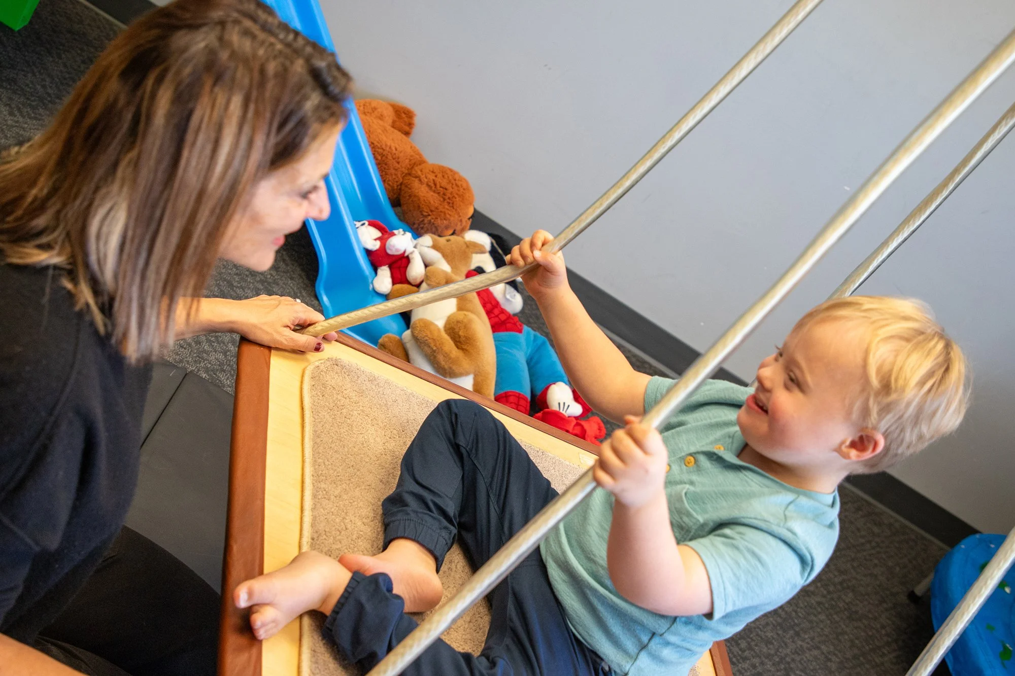 A young boy with blonde hair smiling and playing with an adult woman, possibly his mother, while sitting in a therapy or playroom. The boy holds onto metal bars above a small wooden table with a beige mat on it. There are plush toys, including bears and characters, on a blue slide behind them.