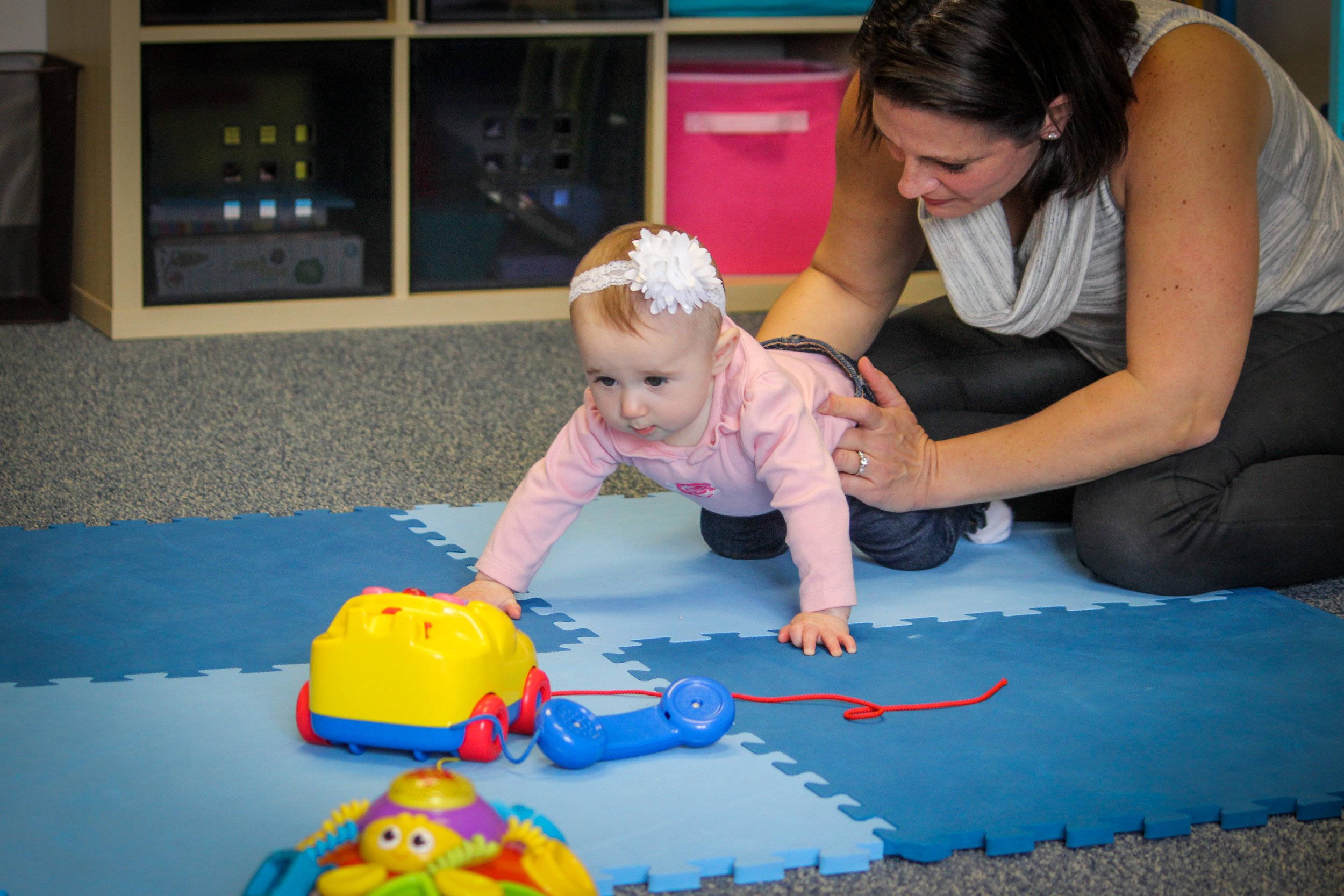 A young child crawling on a blue mat with a woman assisting her. The child is playing with colorful toys, including a toy phone and a yellow toy truck. The woman is on her knees, closely watching and supporting the child.