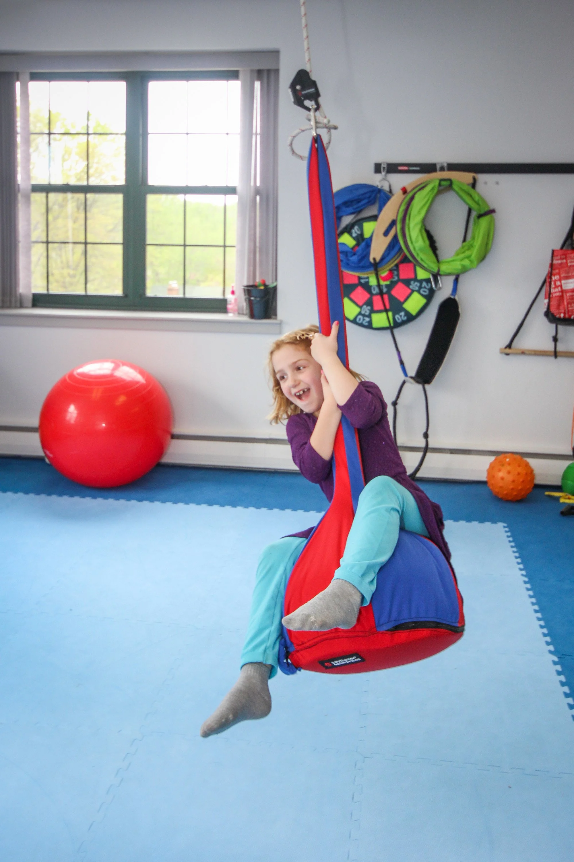 A young girl is smiling and playing on a red and blue hanging swing seat in a playroom with blue foam flooring, gym equipment, and large window showing trees outside.