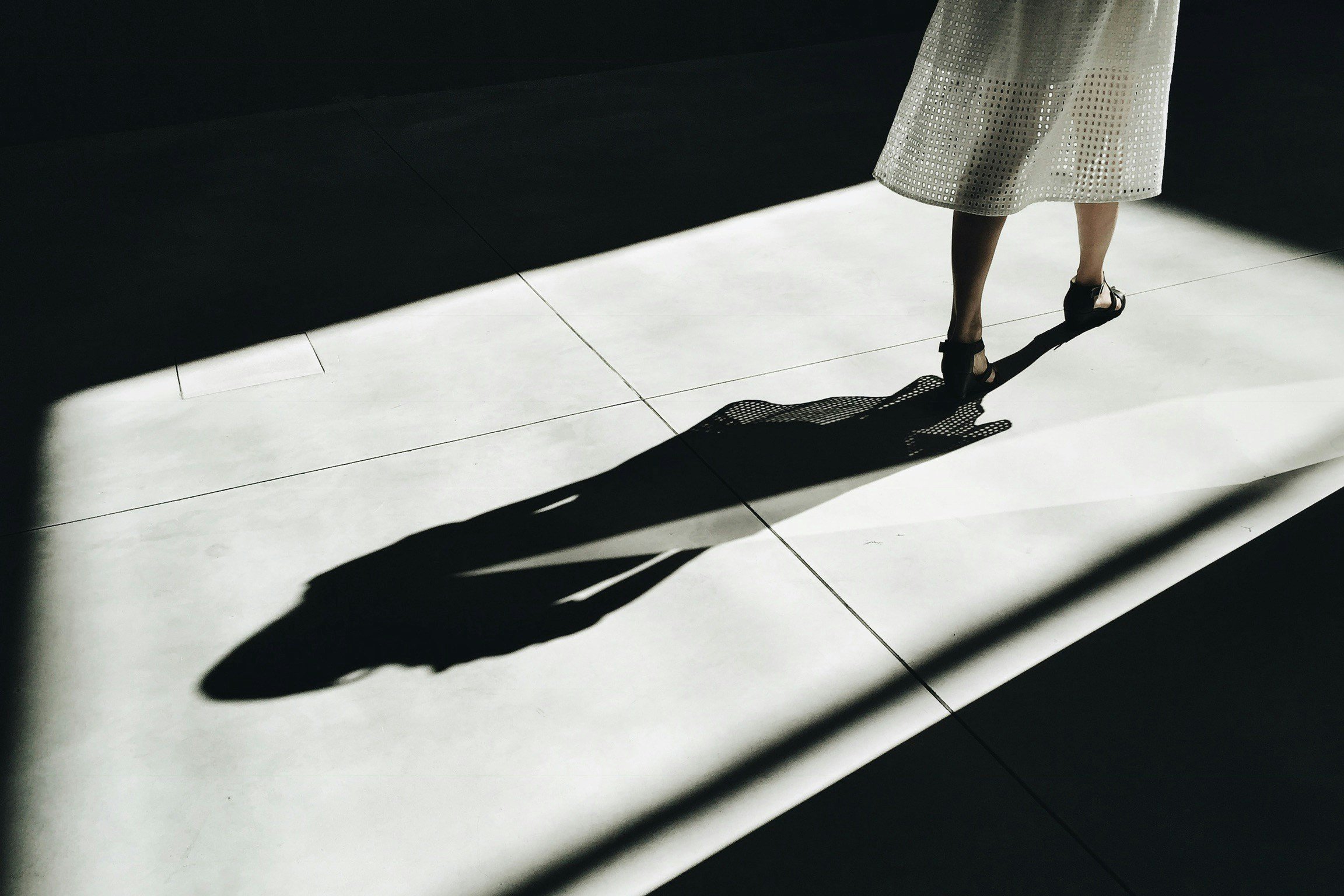 A woman in a patterned skirt and heels standing on a tiled floor, with sunlight casting her shadow on the floor.