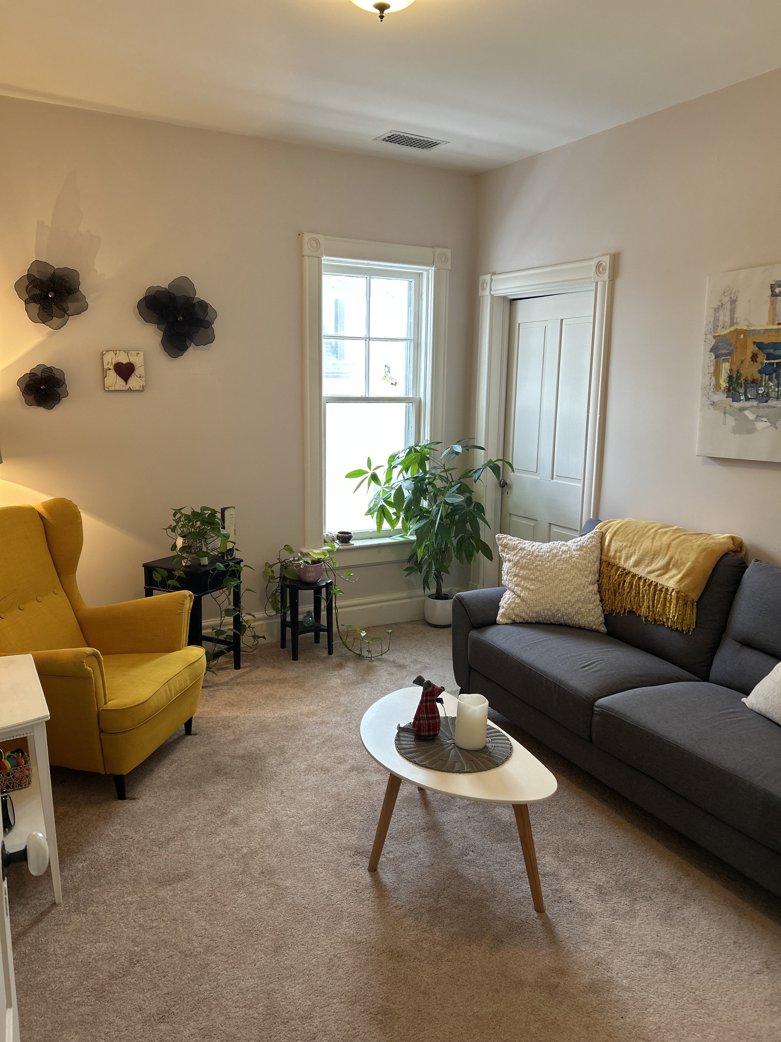 Living room with yellow armchair, gray sofa with pillows, small white coffee table with decorative items, potted plants, a window with curtains, and wall art, organized neatly with carpeted floor.