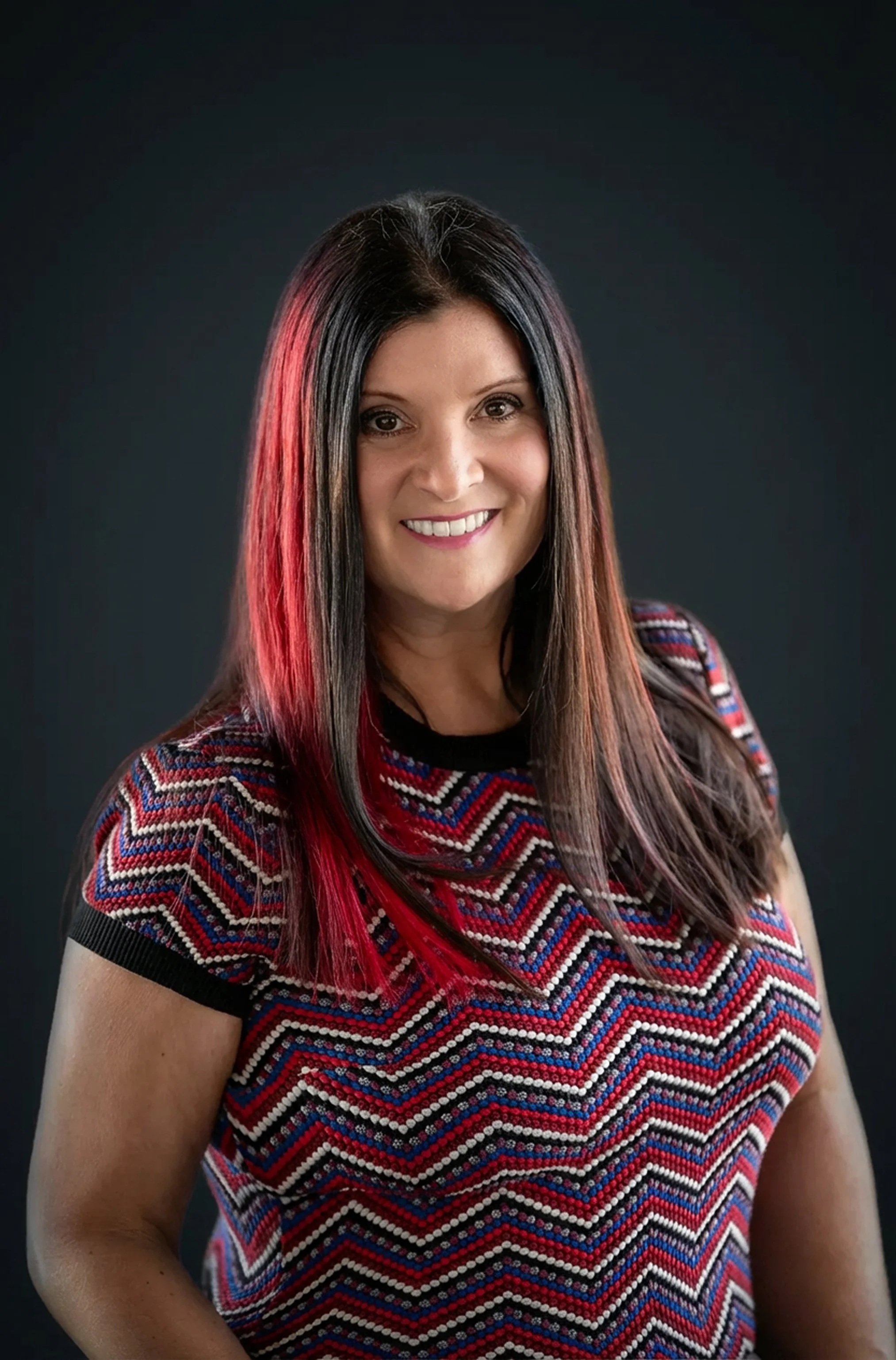 Photo of Dana Cole. A portrait of  a woman with long dark hair accented by red highlights, smiling, against a solid dark background.