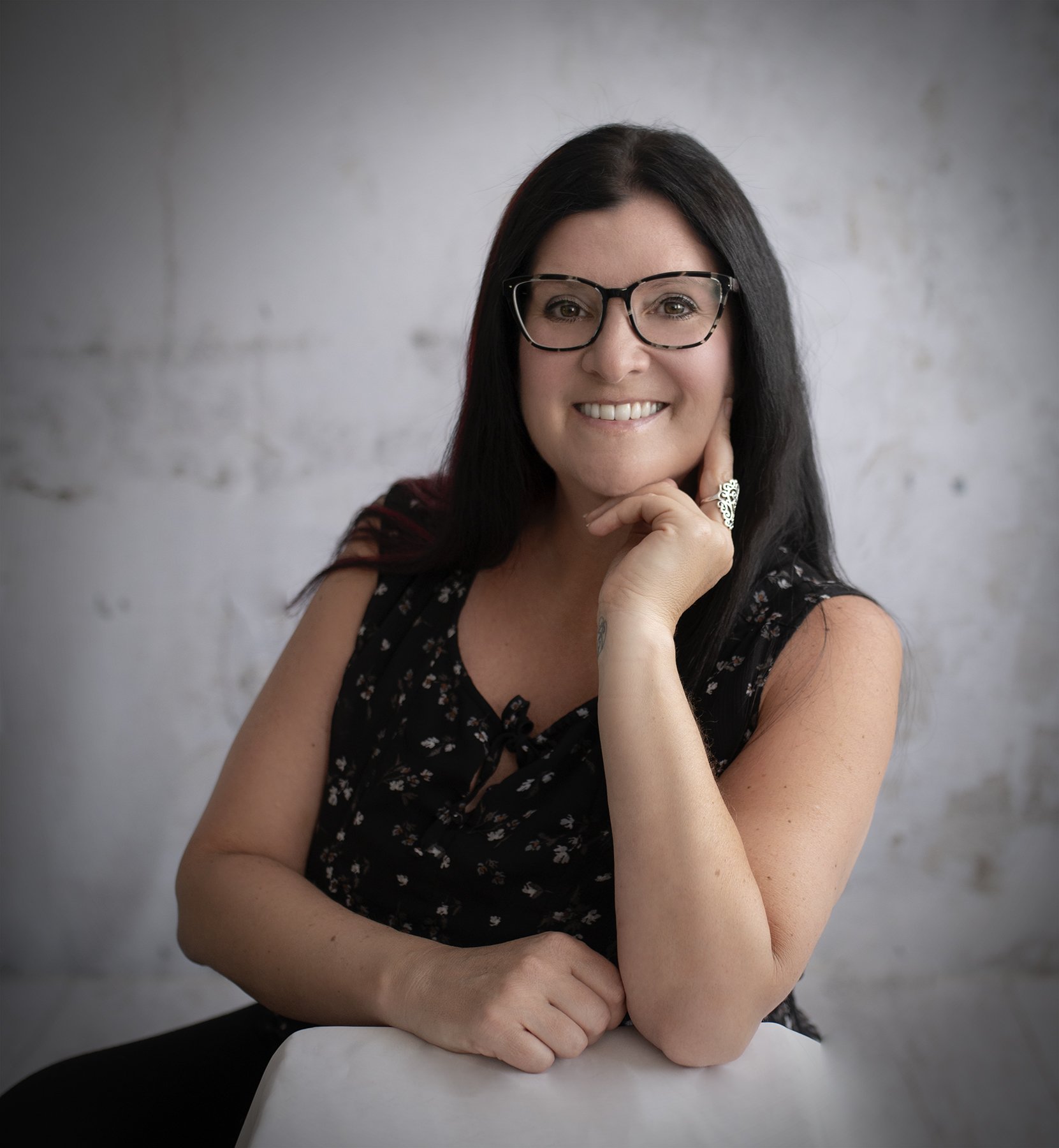 Photo of Dana Cole Coaching, with dark hair and glasses smiling, sitting at a table against a light-colored wall.