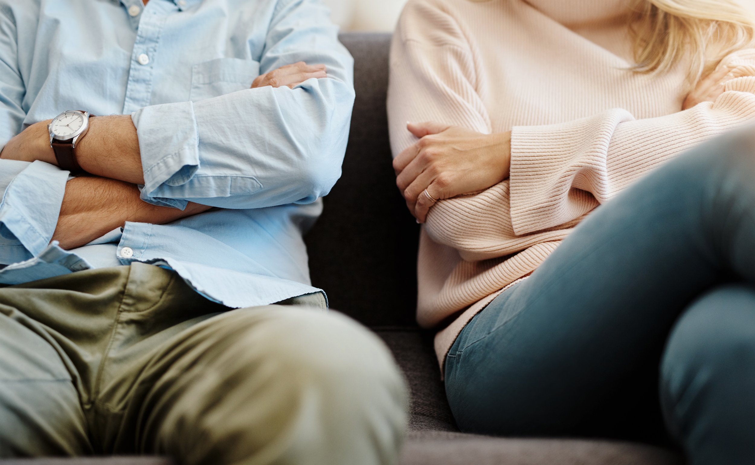 Close-up of two people sitting on a couch with crossed arms, unhappy. One person is wearing a light blue shirt and beige pants, the other is wearing a beige sweater and blue jeans.