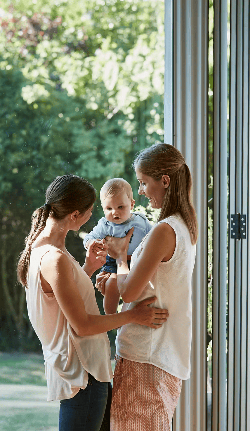 Two women smiling and playing with a baby boy near a large window with greenery outside.