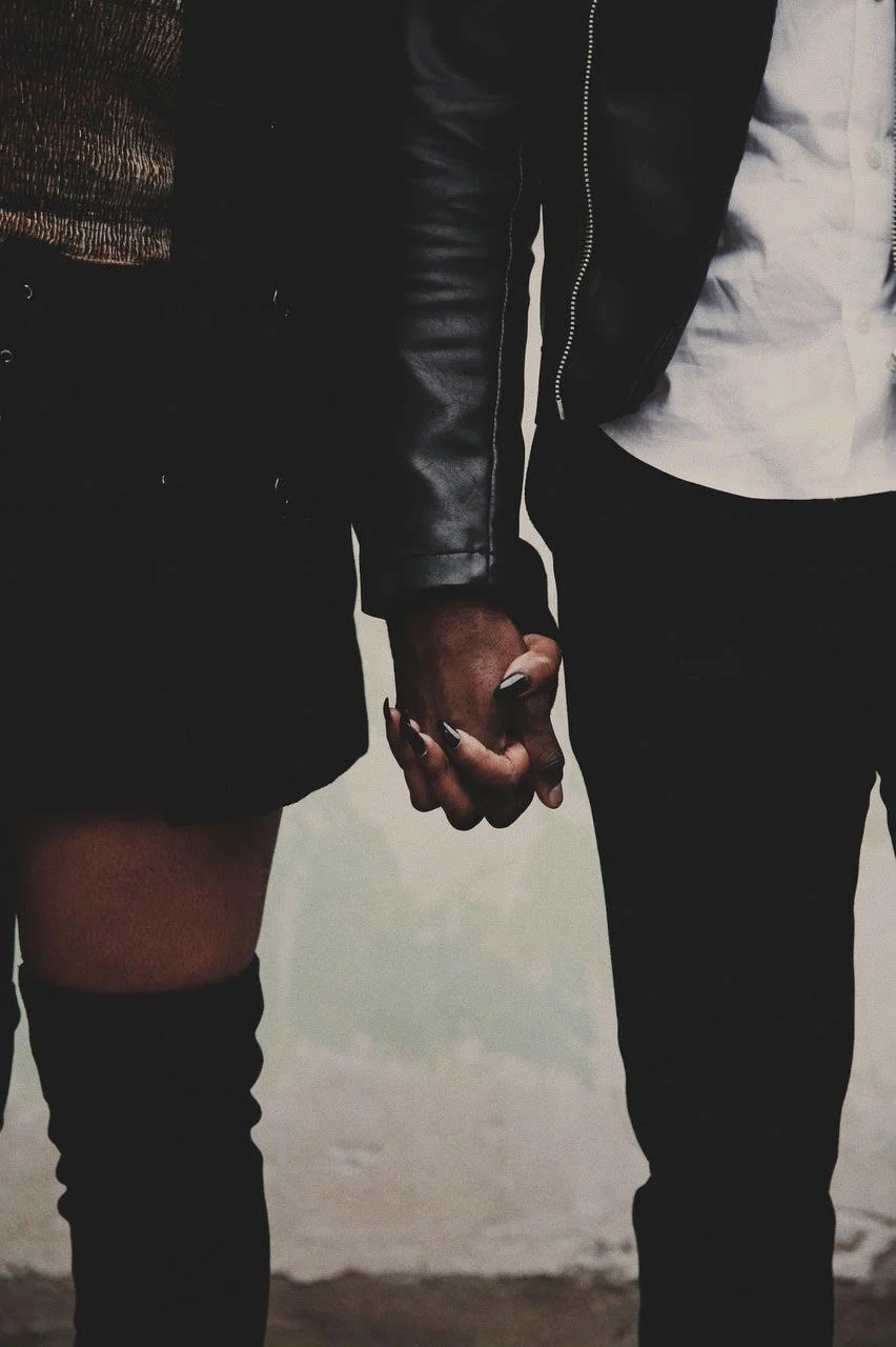 Close-up of two people holding hands, one person wearing a black leather jacket and the other in a white shirt, with a blurred background.