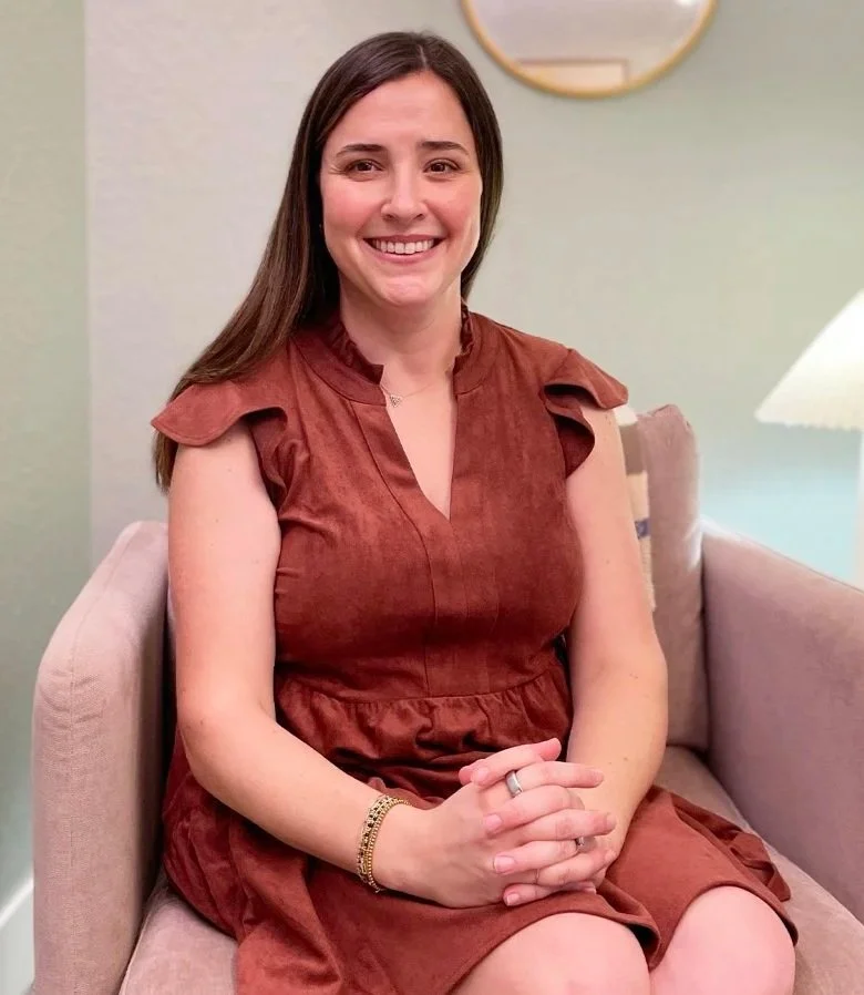 A smiling woman with long brown hair, wearing a rust-colored dress with ruffle sleeves, sitting on a pink armchair in a cozy room with a lamp, mirror, and light-colored walls.