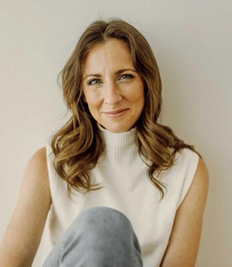 A woman with wavy brown hair wearing a white sleeveless turtleneck shirt, smiling and sitting against a plain light-colored wall.