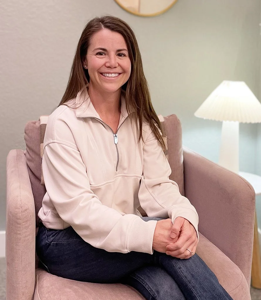 A woman with long brown hair smiling while sitting on a pink armchair in a cozy living room. She is wearing a light-colored zip-up jacket and dark jeans, with a white lamp and a decorative wall clock in the background.