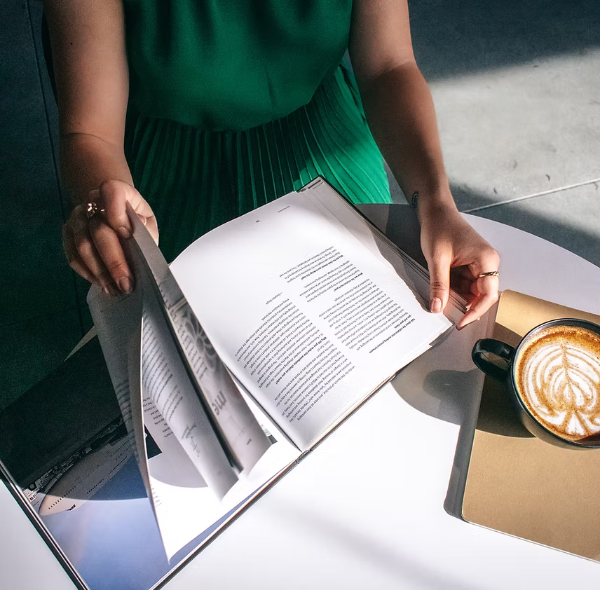 Person wearing a green dress flipping through pages of a book or magazine at a table with a latte in a black mug on a gold tray.
