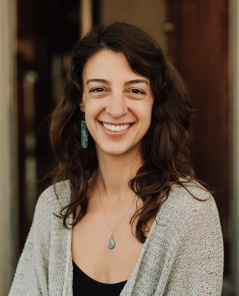 A smiling woman with dark, curly hair wearing a grey cardigan, turquoise earring, and a pendant necklace, standing outdoors with a blurred wooden background.