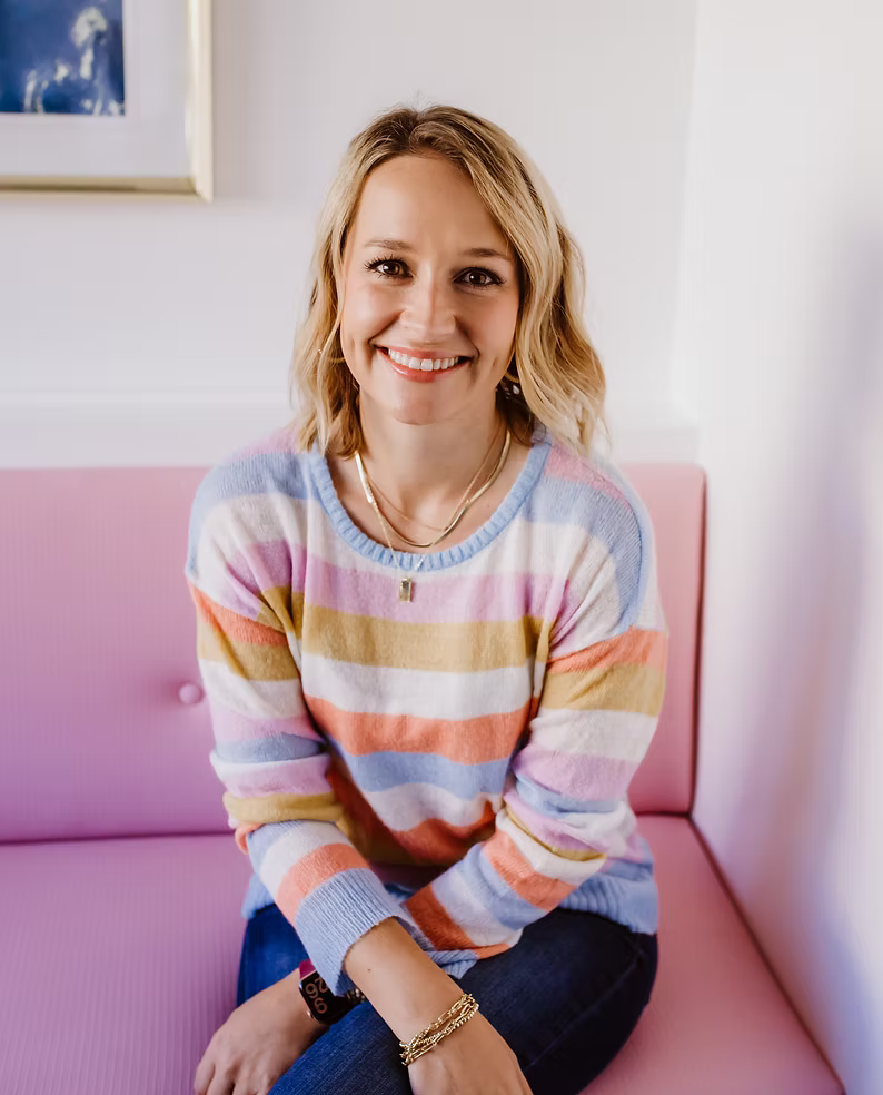 A smiling woman with shoulder-length blonde hair sitting on a pink bench, wearing a pastel rainbow-striped sweater, jeans, and jewelry in a bright, white room.